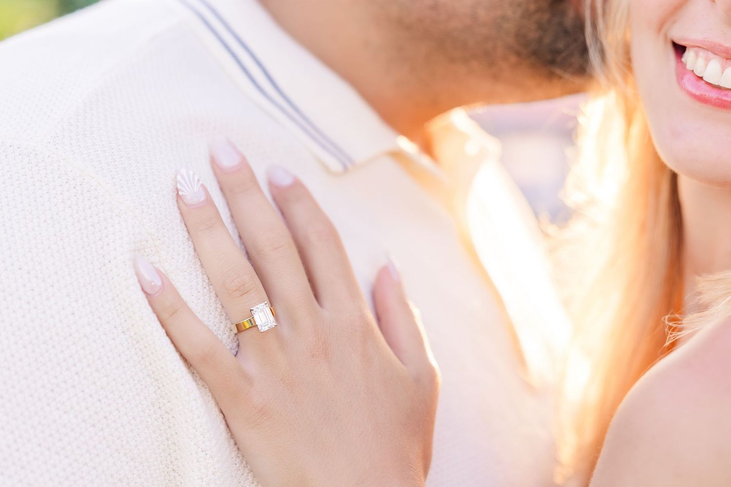 Close-up of a woman's hand showing a gold engagement ring while embracing her partner in sunlight.