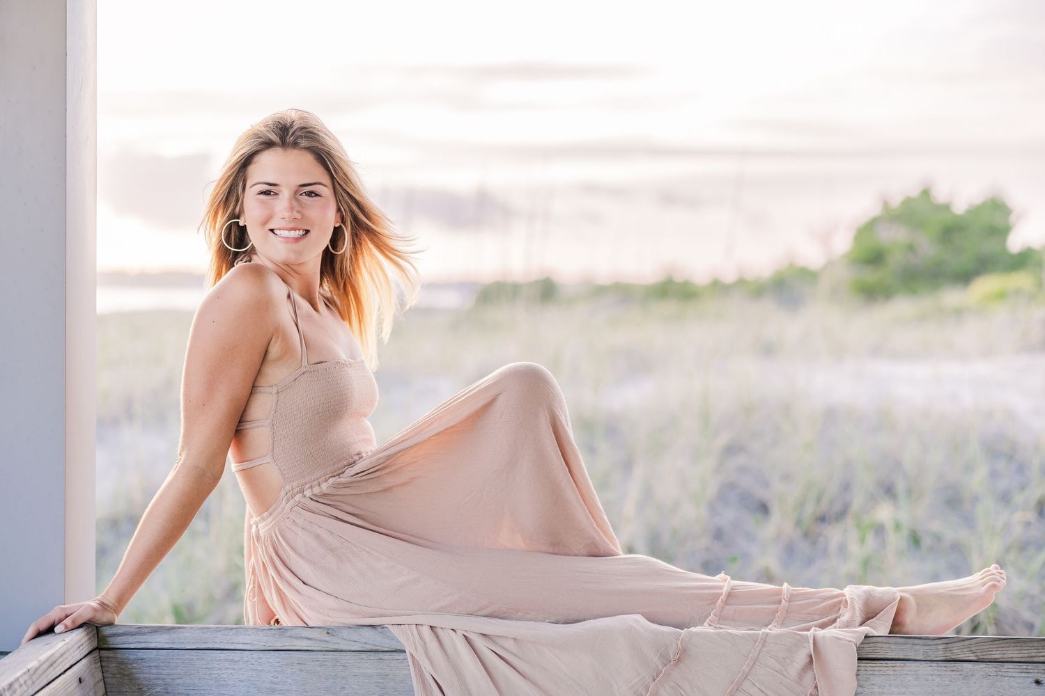 A dreamy beach photoshoot featuring a flowing mauve dress against sand dunes and wooden fencing at sunset.
