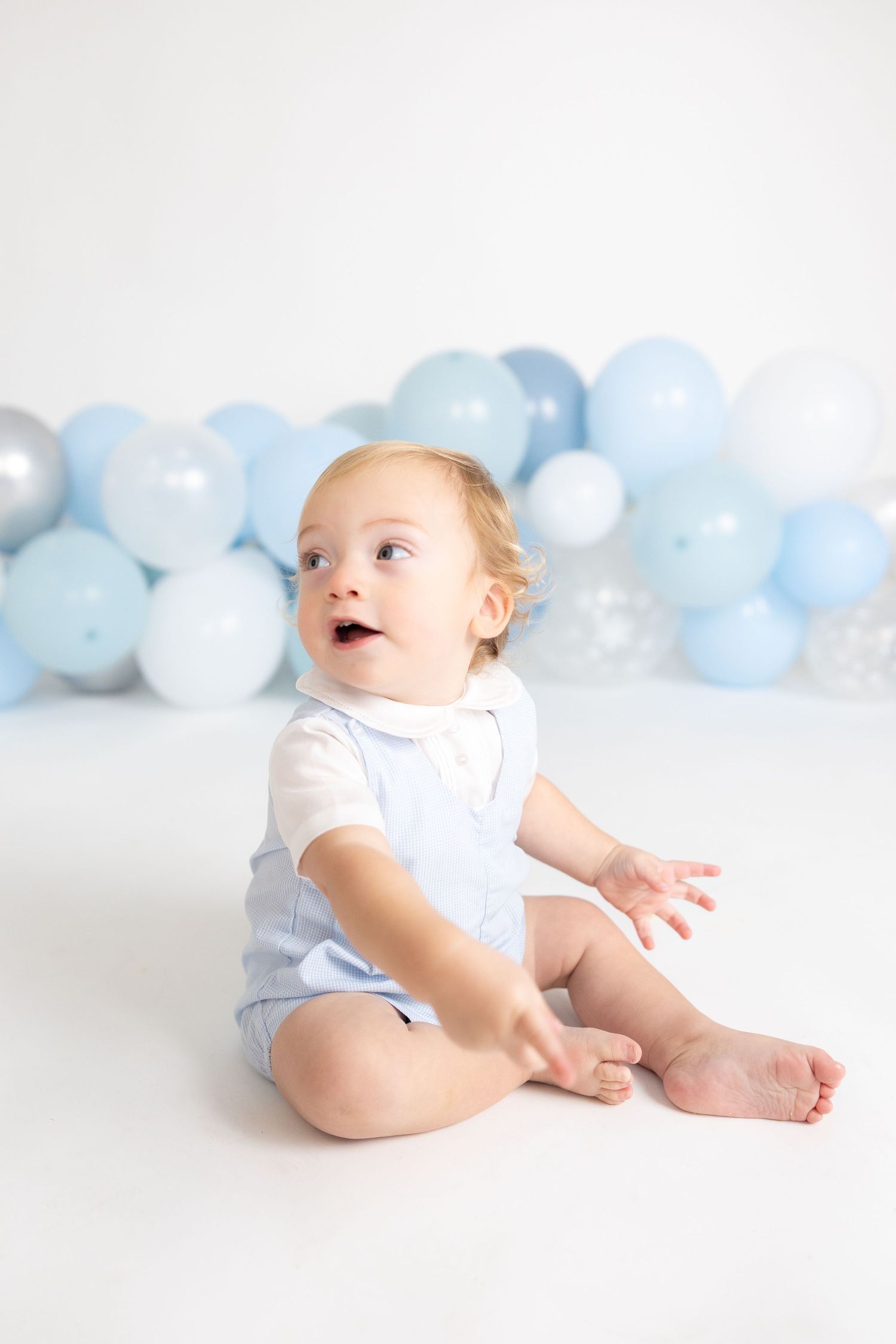 Baby in white outfit playing on floor with blue and silver balloon display.