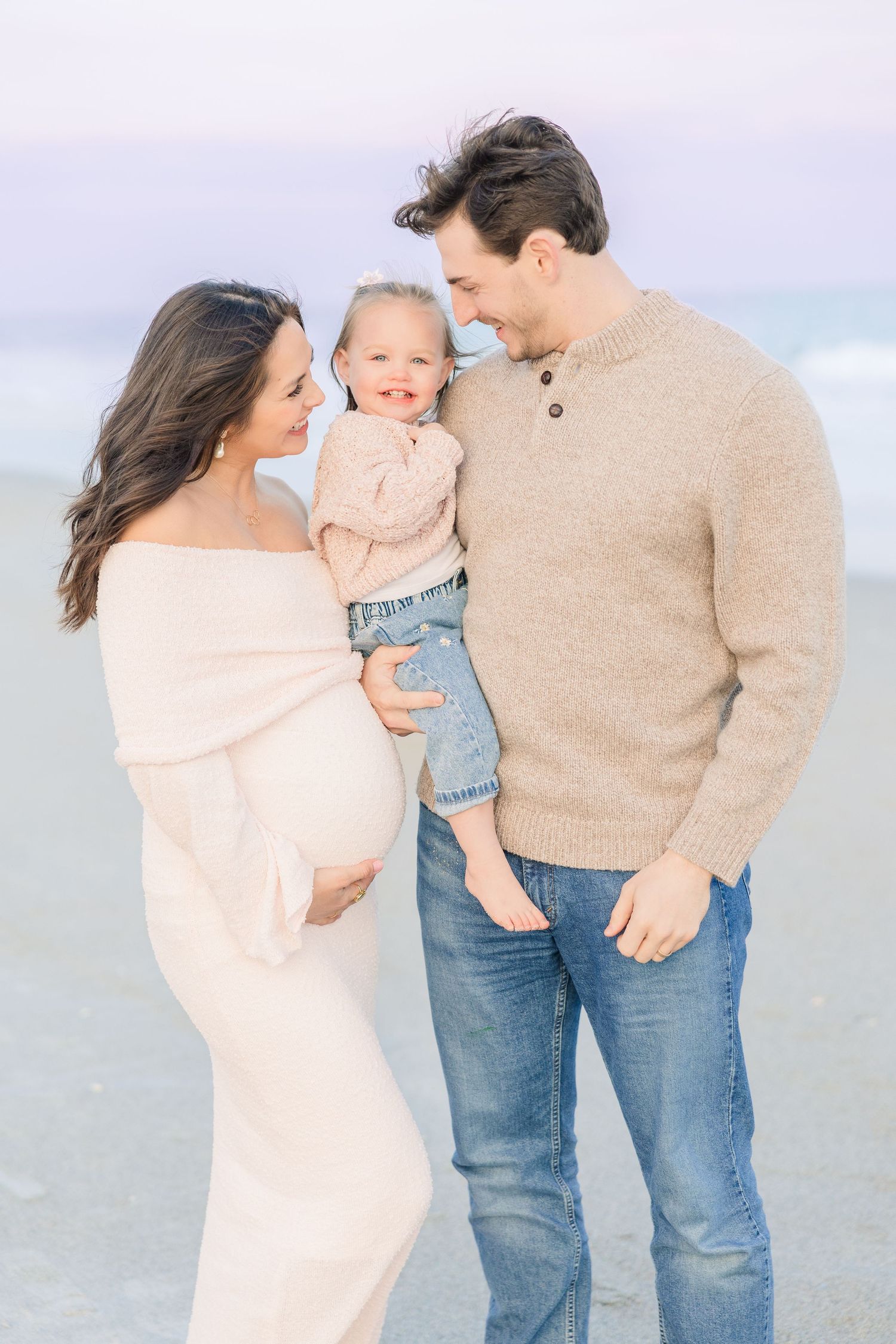 Family shares tender moments together on a cloudy beach in neutral colored clothing.