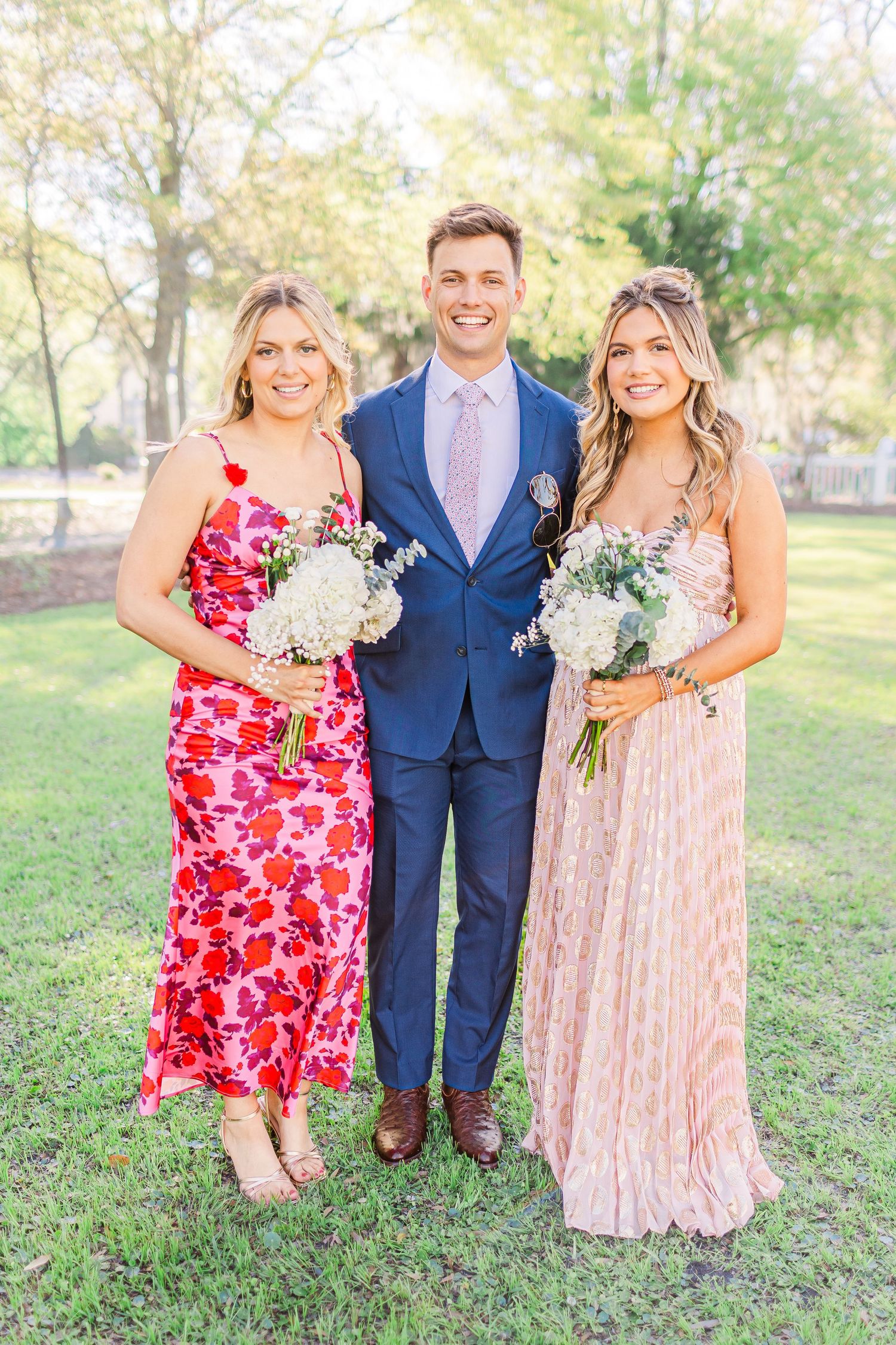 Wedding party members in floral dresses and navy suits posing together on grass.