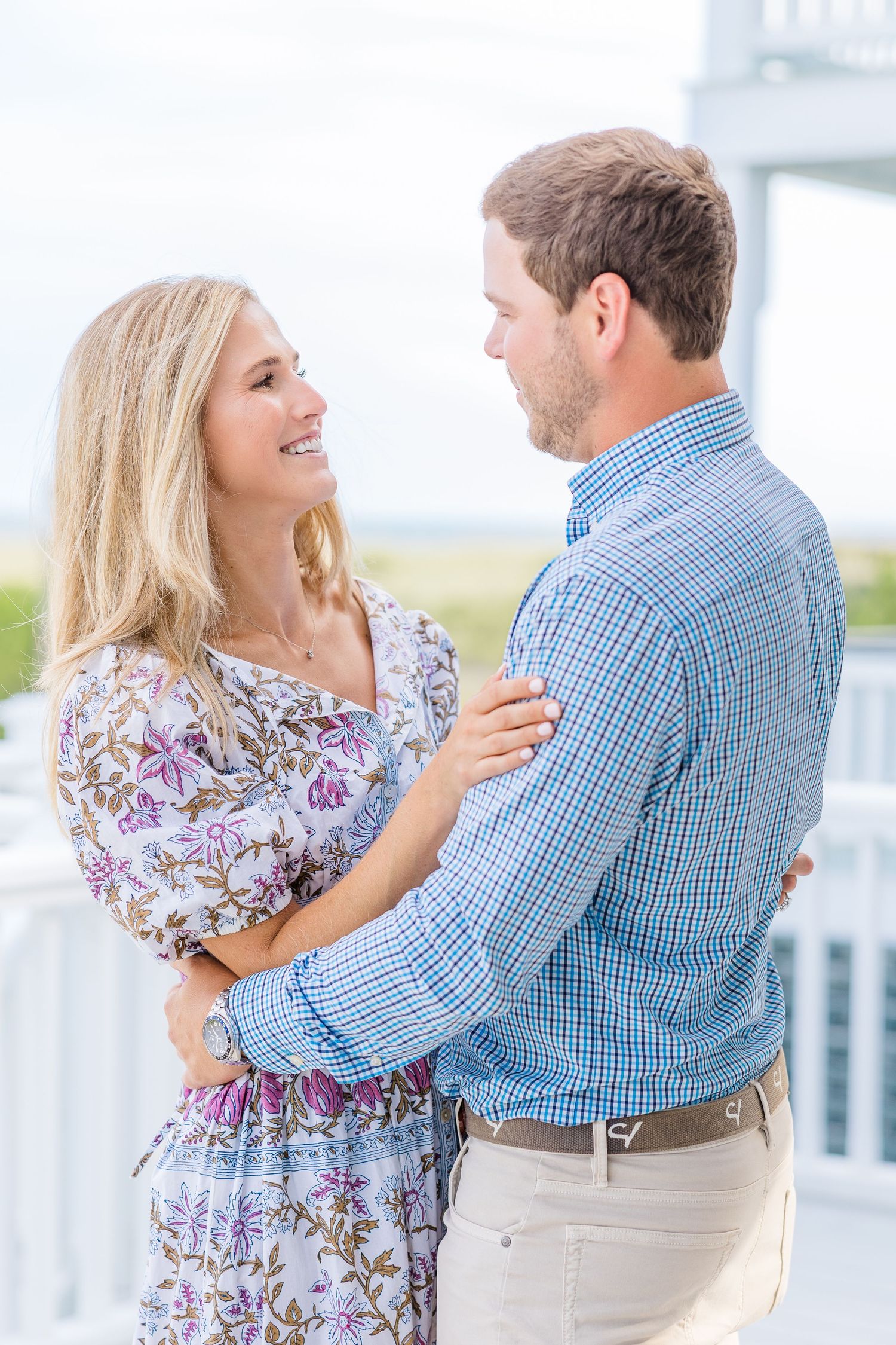 Happy couple in summer attire embracing on a white porch with coastal views.