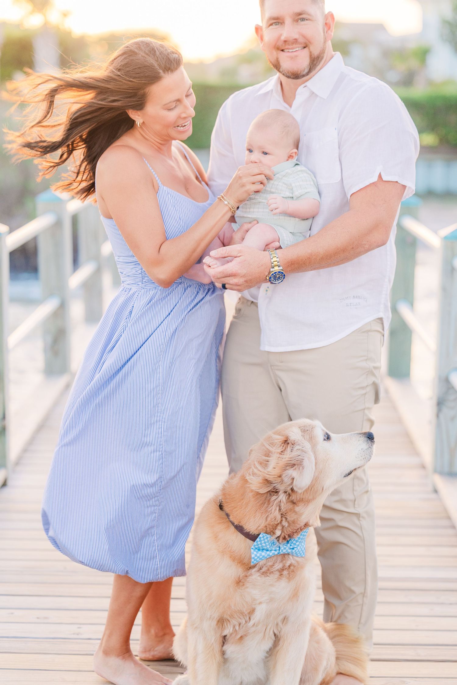 A golden retriever with a blue bowtie stands with a family on a wooden boardwalk at sunset.