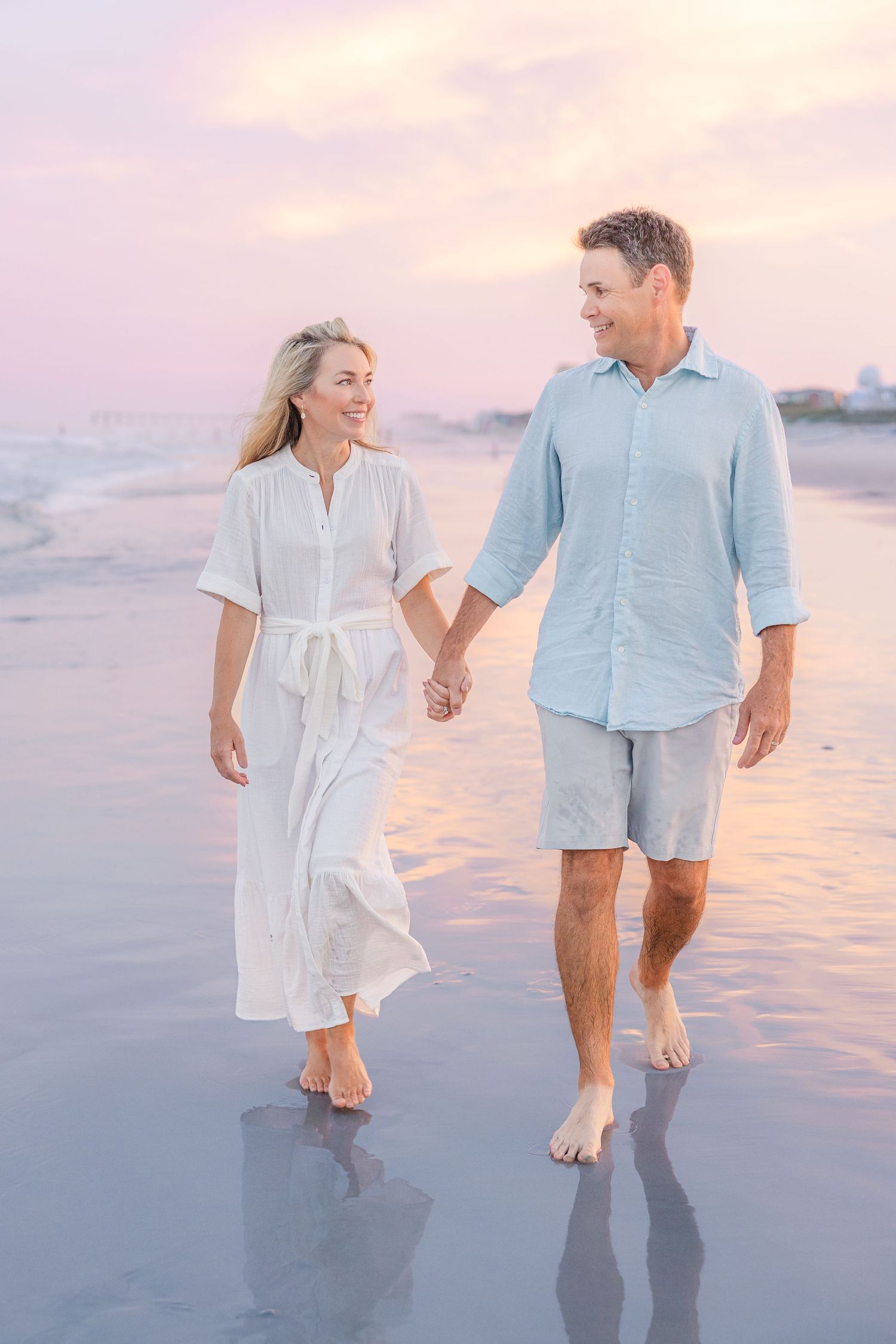 A romantic beach walk at sunset with a couple in light summer clothing holding hands.