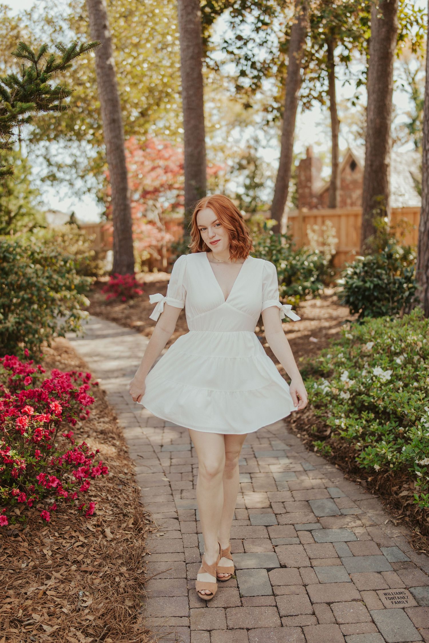 Red-haired woman in a white dress walking down a garden path lined with vibrant flowers during a spring portrait session in Wichita, Kansas,