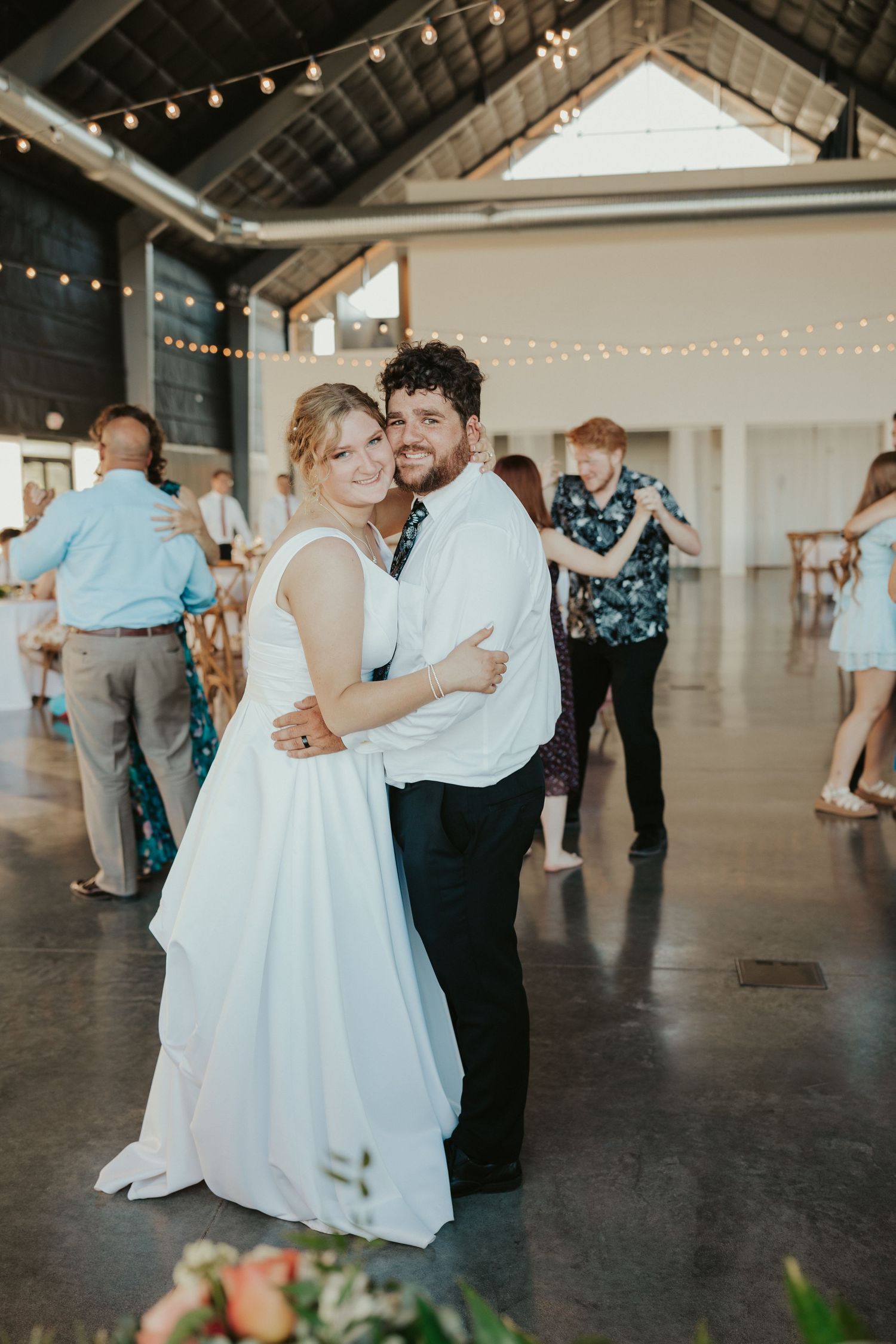 Joyful bride and groom dancing at their wedding reception under string lights, surrounded by guests, in a Kansas wedding venue