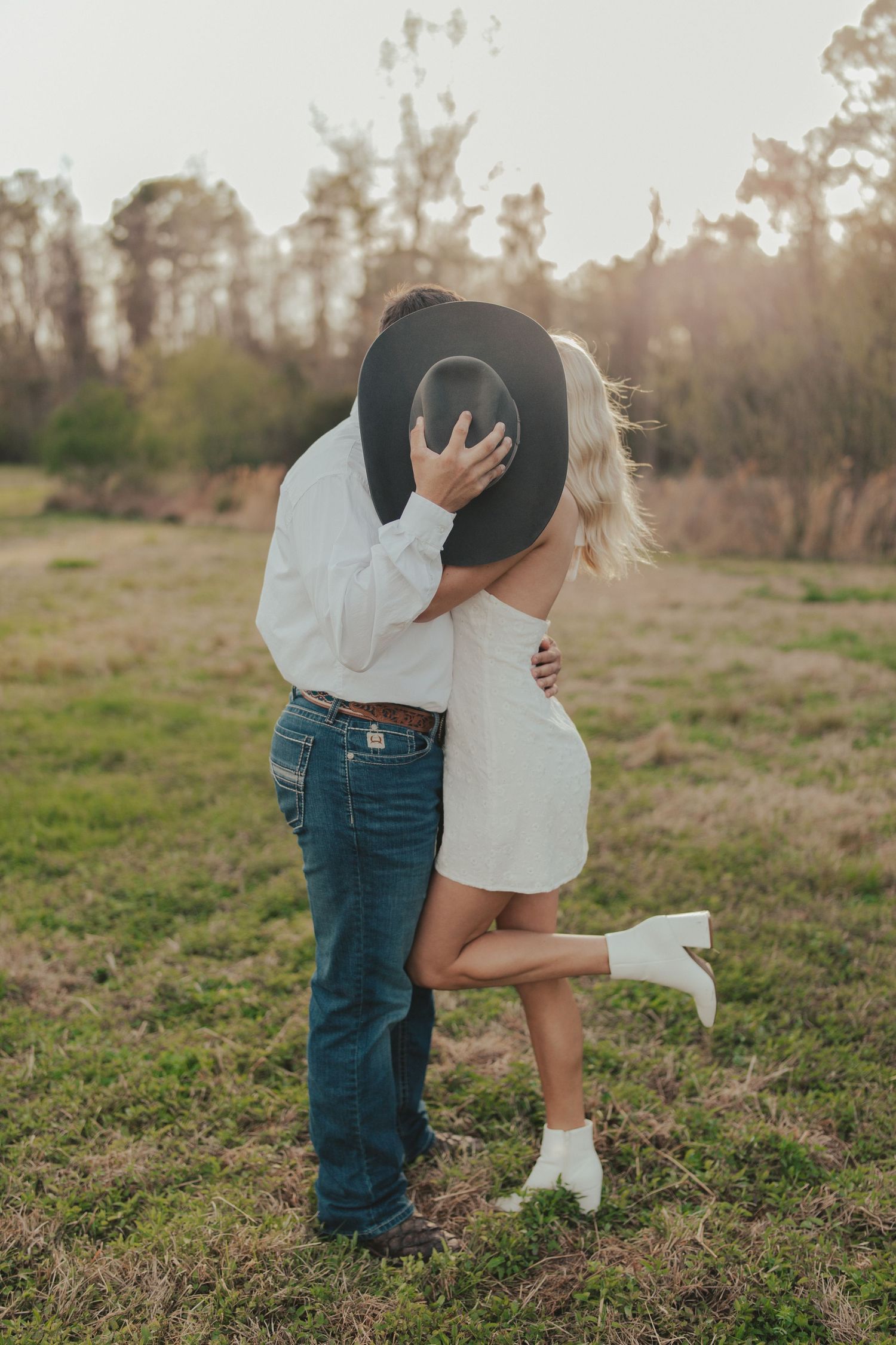 Engagement photo of a couple standing in a sunlit field in Kansas, with the man playfully covering their faces with a black cowboy hat