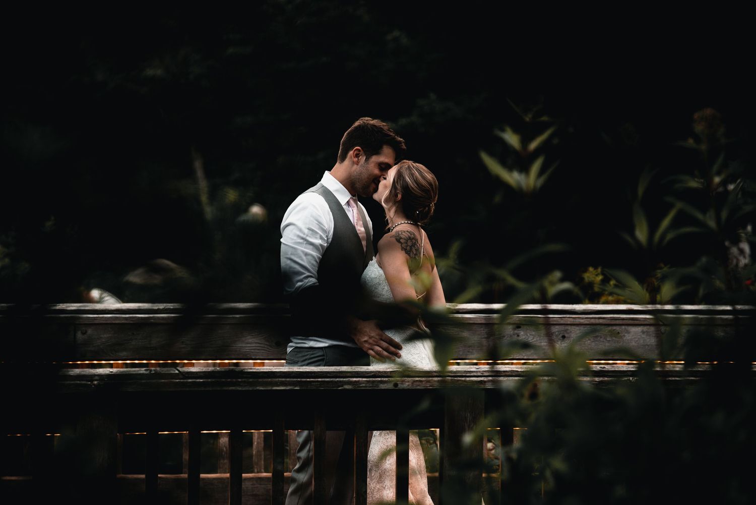Bride and groom embracing on a bridge during an outdoor wedding, photographed through foliage.