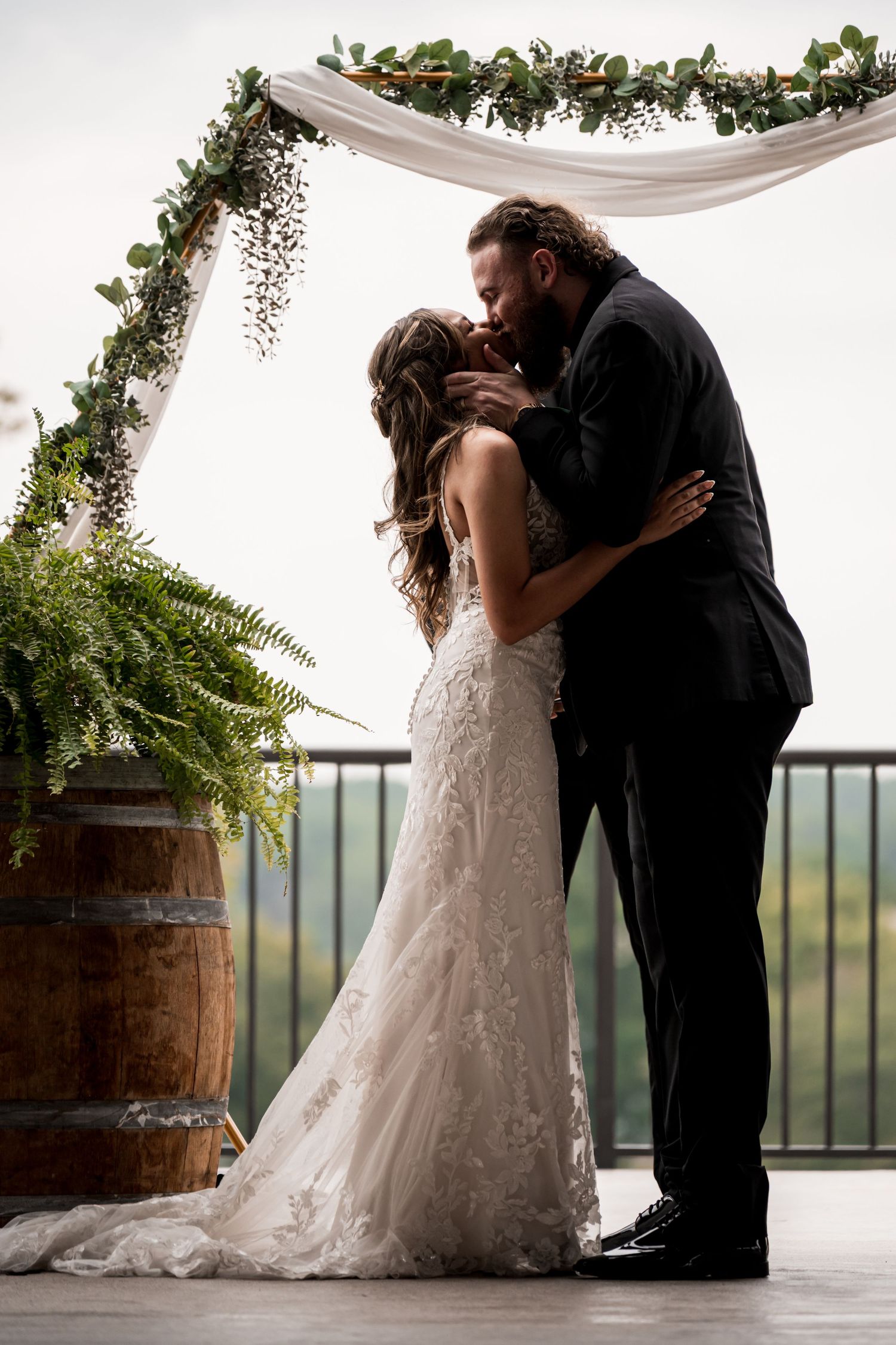 Bride and groom sharing their first kiss beneath a floral arch with wine barrels nearby.