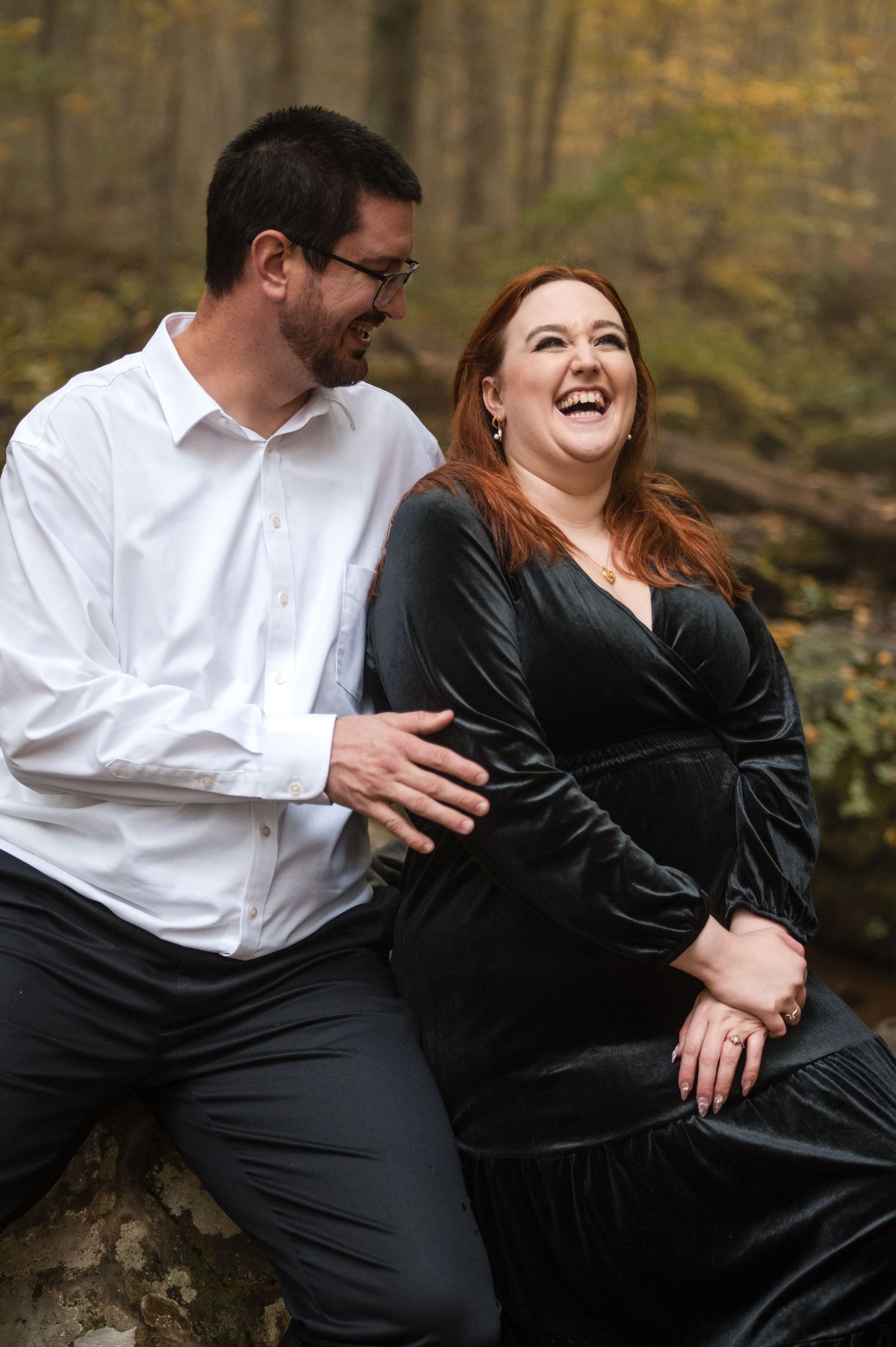 Couple in formal attire laughing together in a fall forest setting.