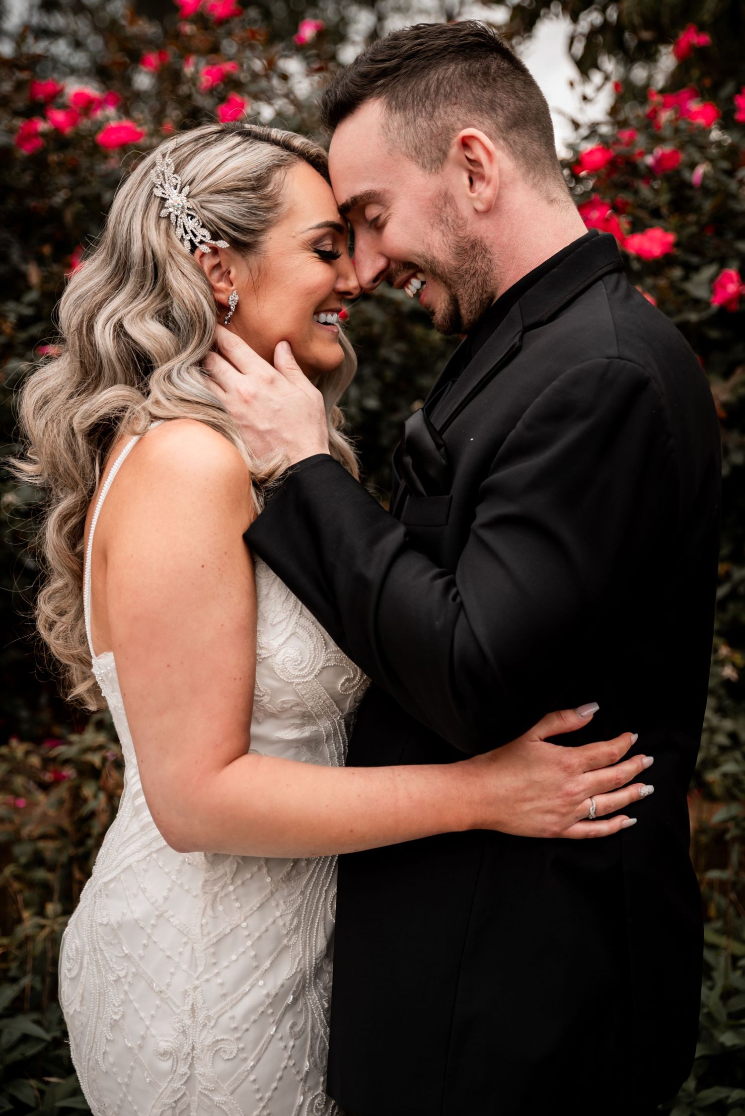 Bride and groom standing together in front of a backdrop of red roses.