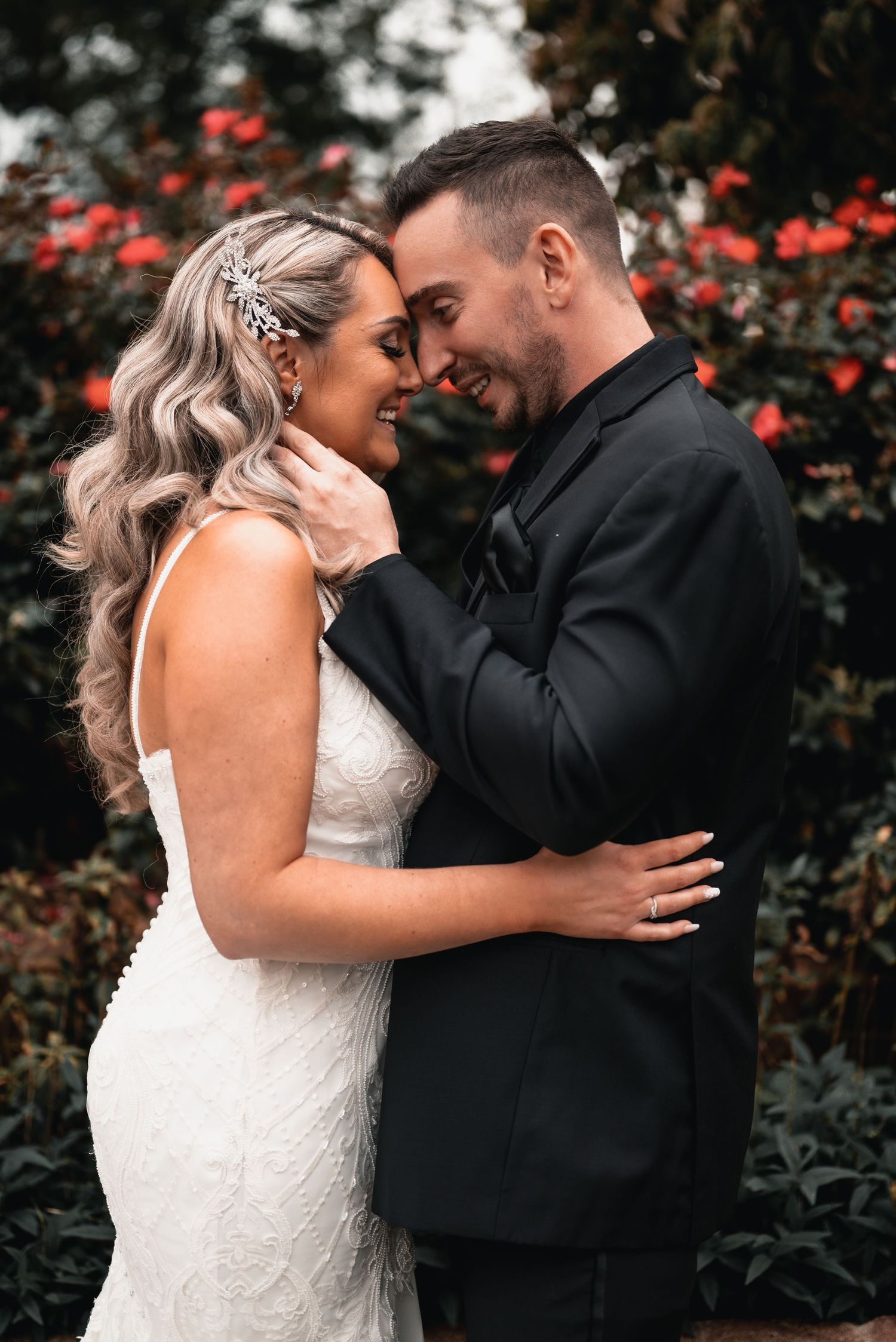 Bride and groom standing together in a garden wedding setting with red flowers behind them.