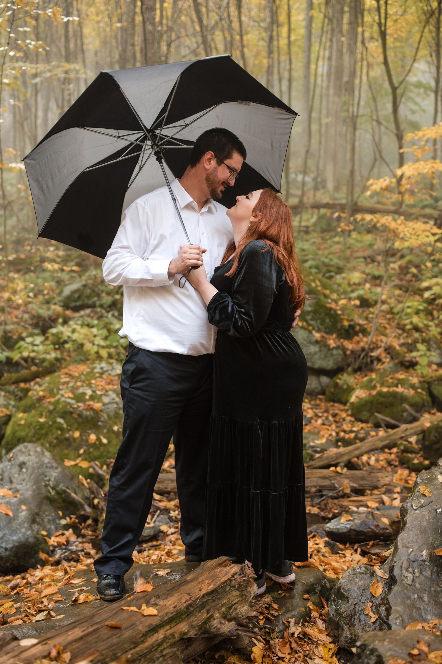 Couple standing together under a black umbrella in an autumn woodland setting.
