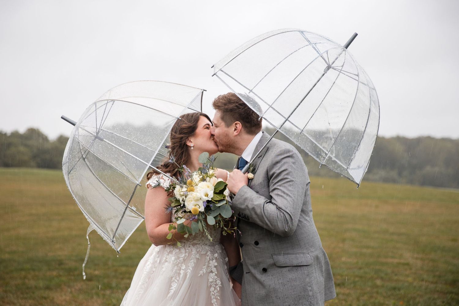 Couple standing together under clear umbrellas in an open field.