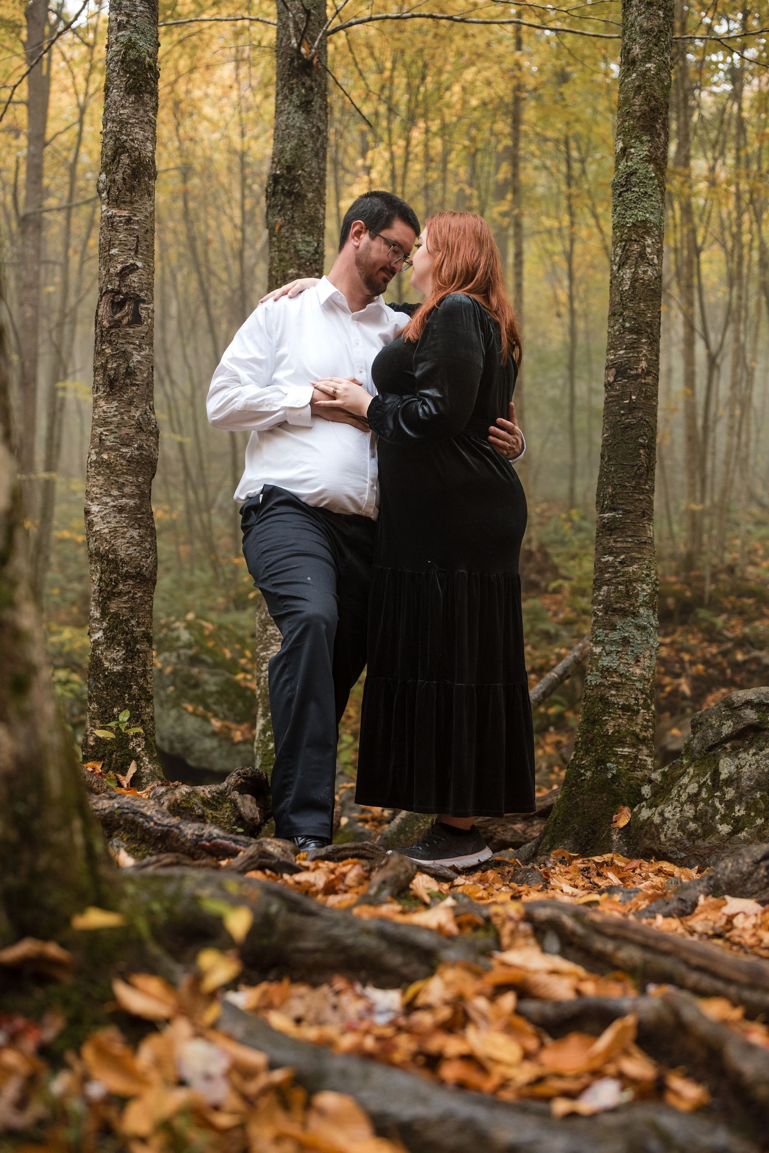 Couple standing together in an autumn forest surrounded by fallen leaves.
