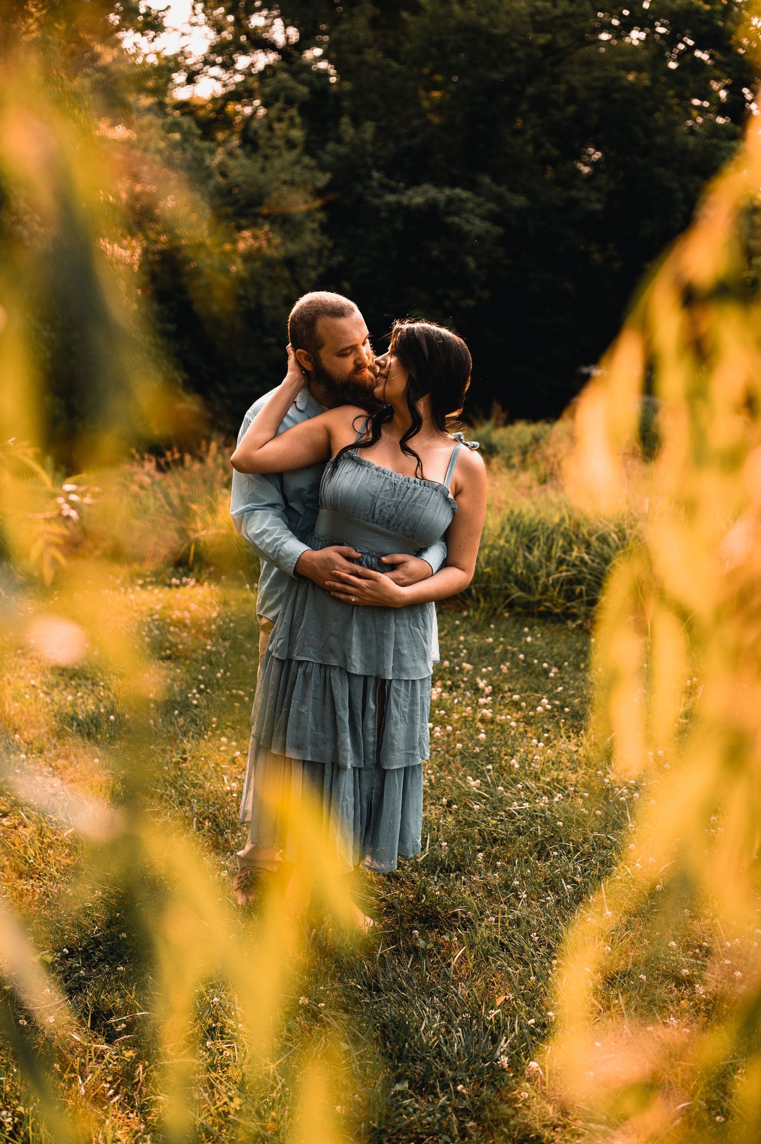 Couple standing together in a sunlit meadow with golden wildflowers in the foreground.