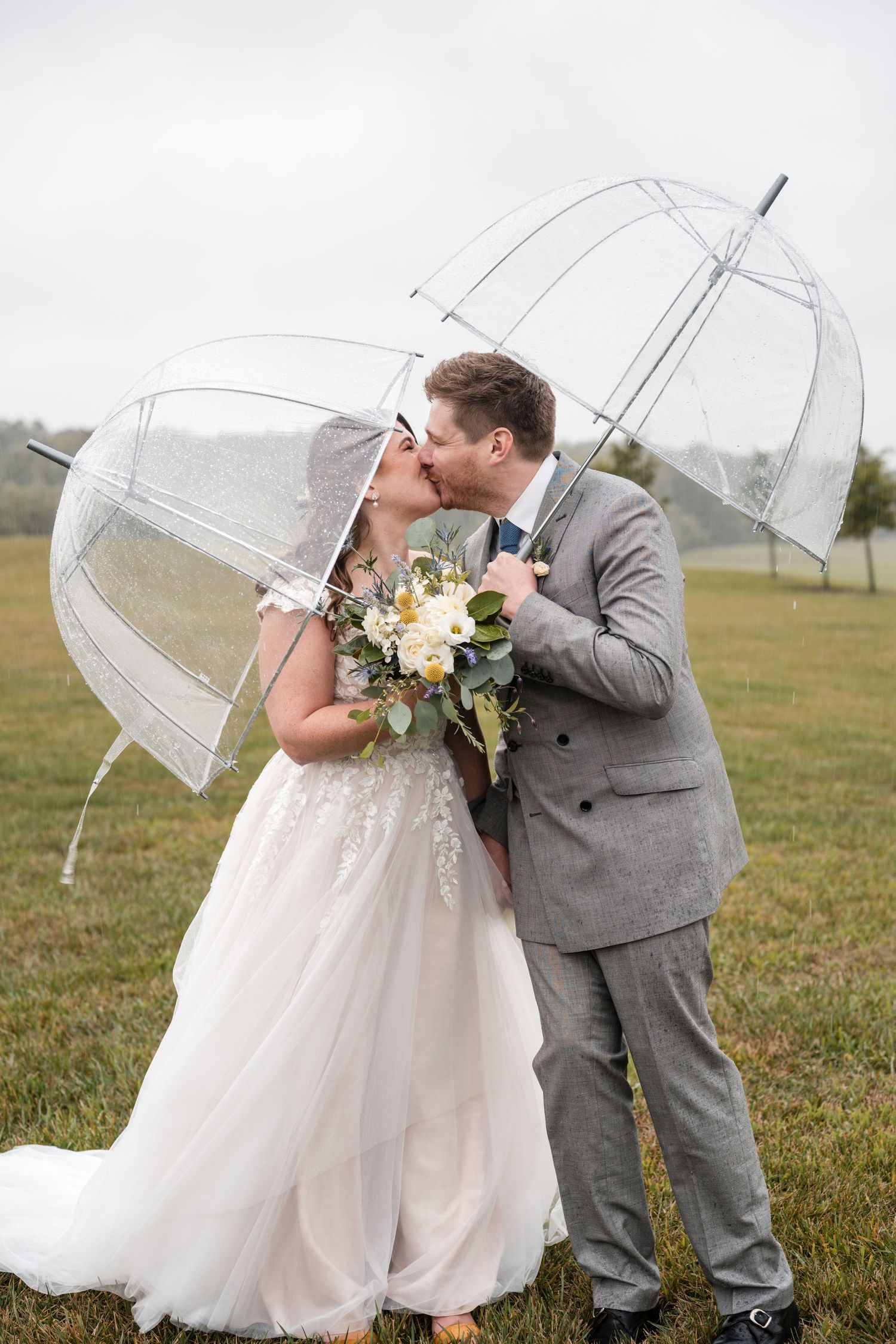 Bride and groom standing together with clear umbrellas in an open field on a rainy wedding day.