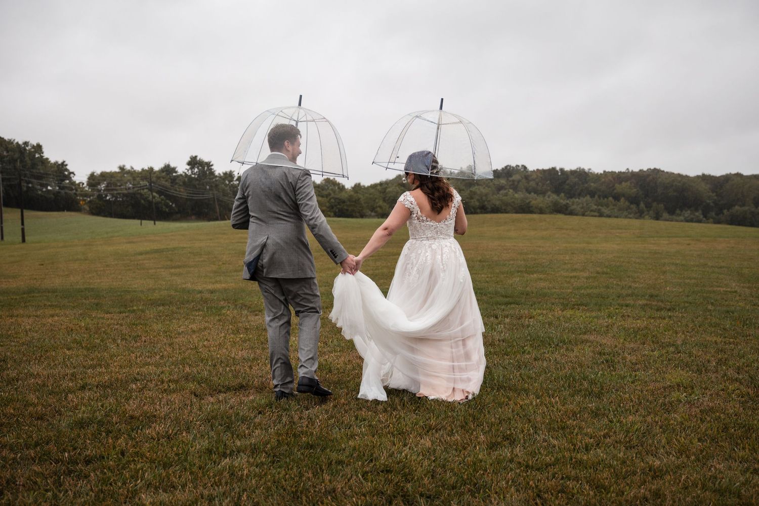 Bride and groom walking together across a grassy field while holding clear umbrellas.