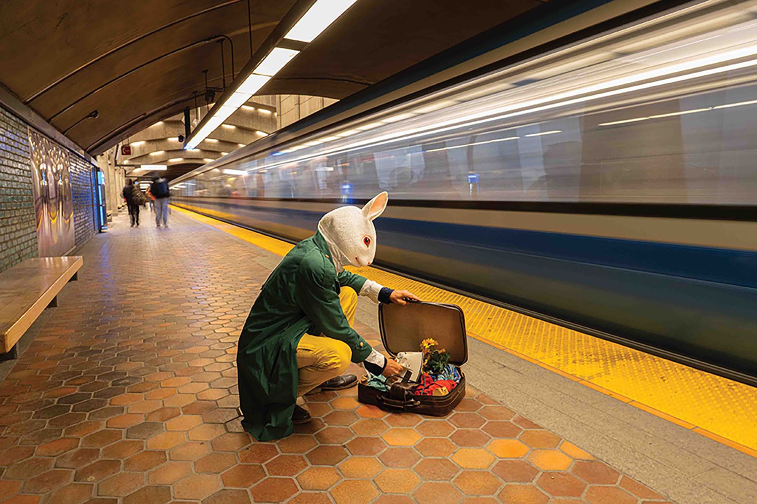 Person in Easter bunny costume crouching with open suitcase on subway platform as train passes by.