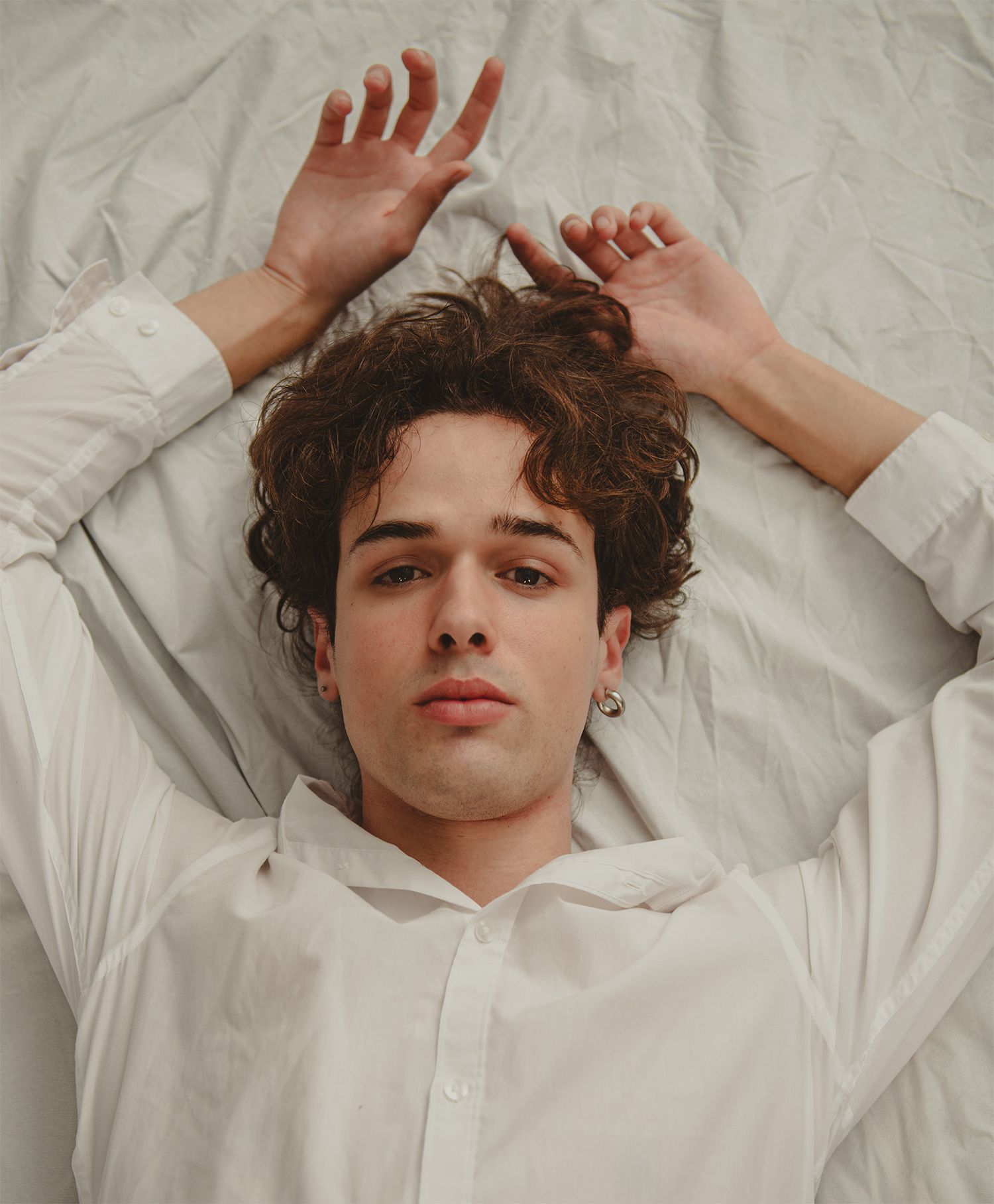 Young man with curly brown hair lying on white bedding, wearing a white shirt, hands raised beside his head.
