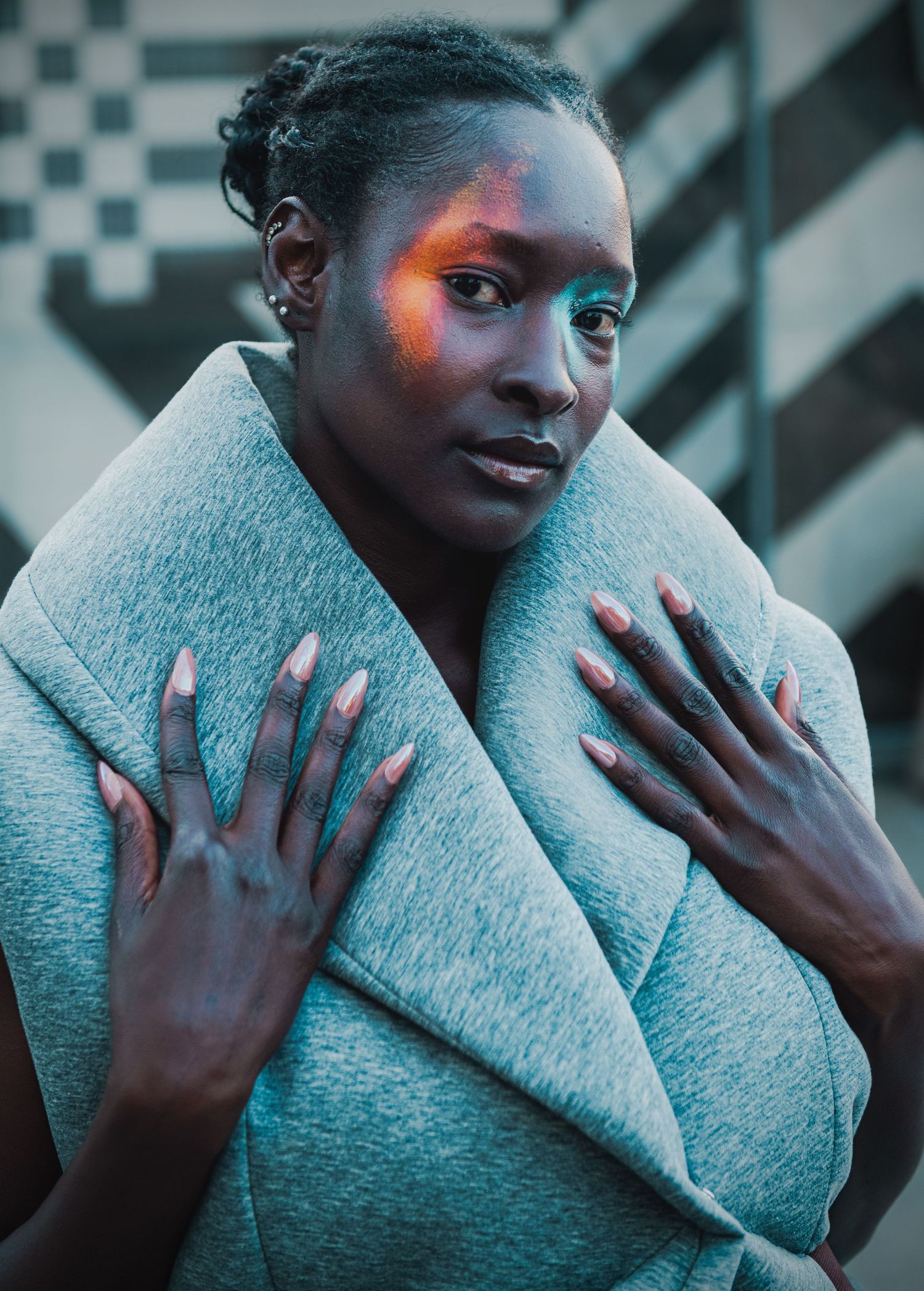 A Black woman with colorful eye makeup poses in a gray wrap, hands crossed over chest, against an urban background.