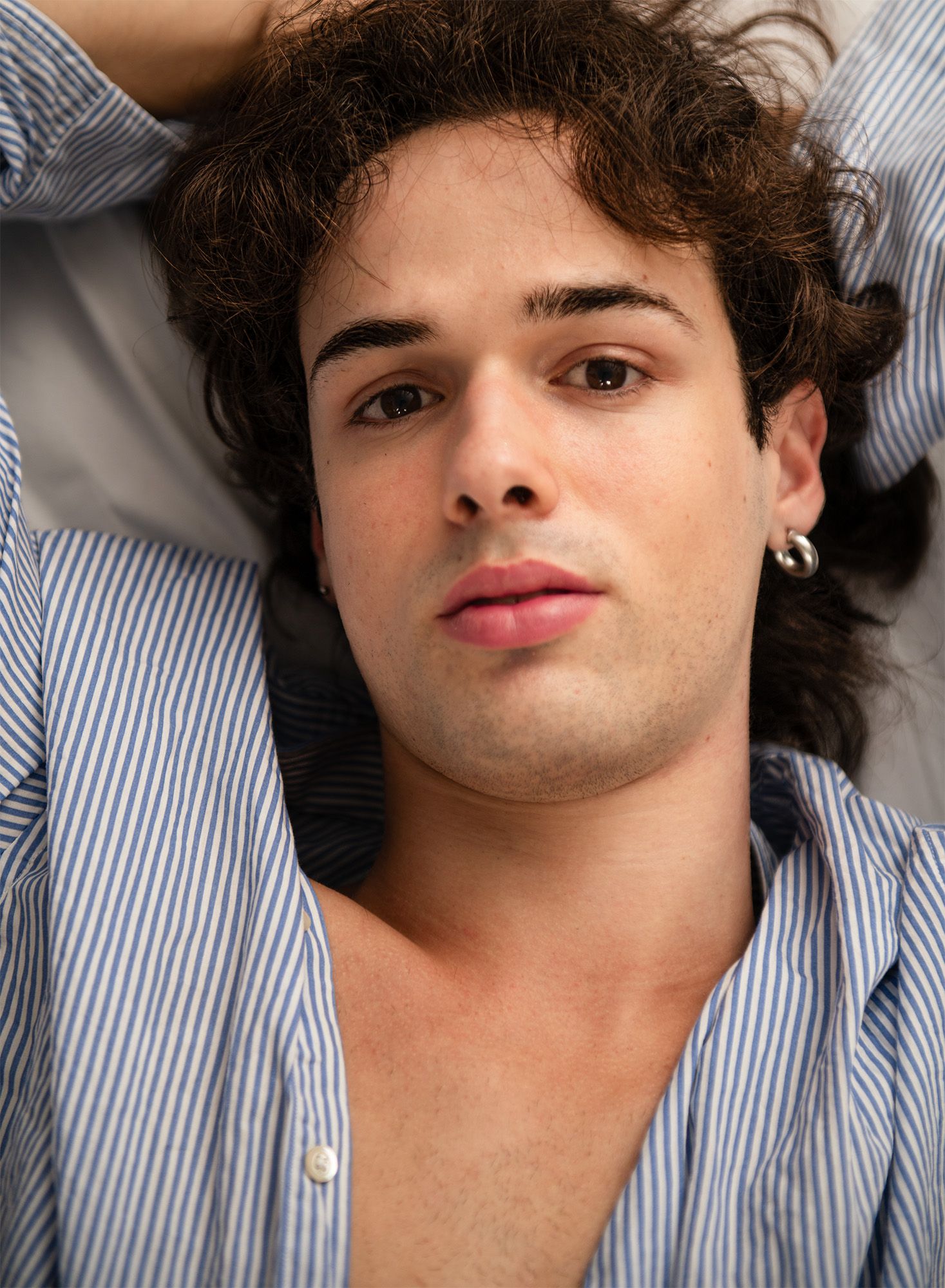 Young man with curly dark hair and hoop earring lying down, wearing an open blue striped shirt.