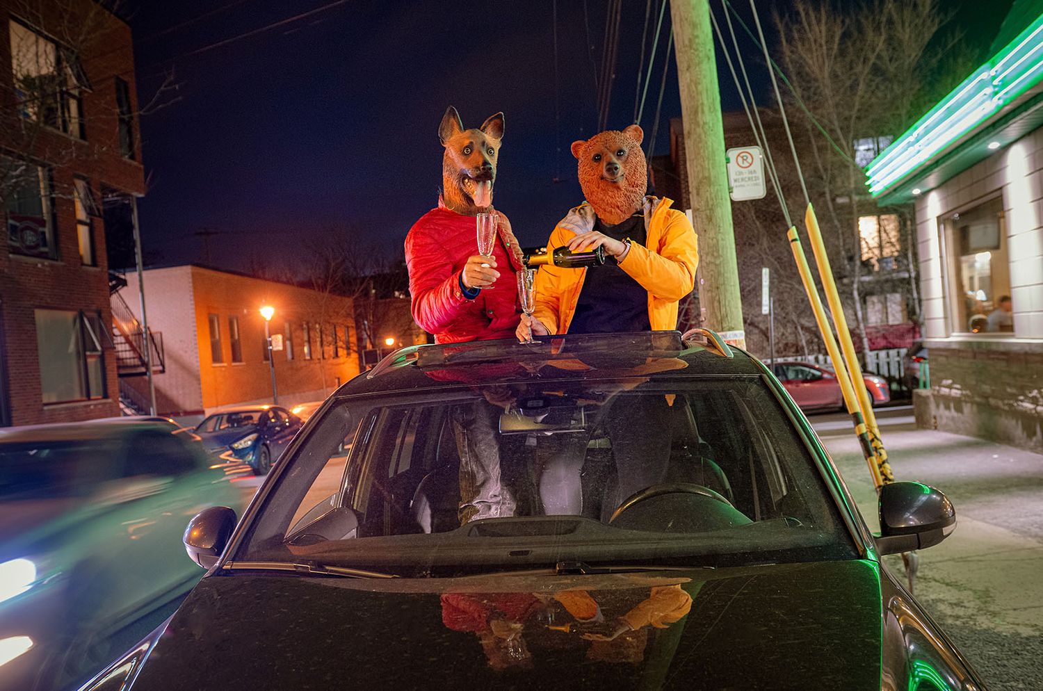 Two people wearing animal masks popping out of a car sunroof at night on a city street.