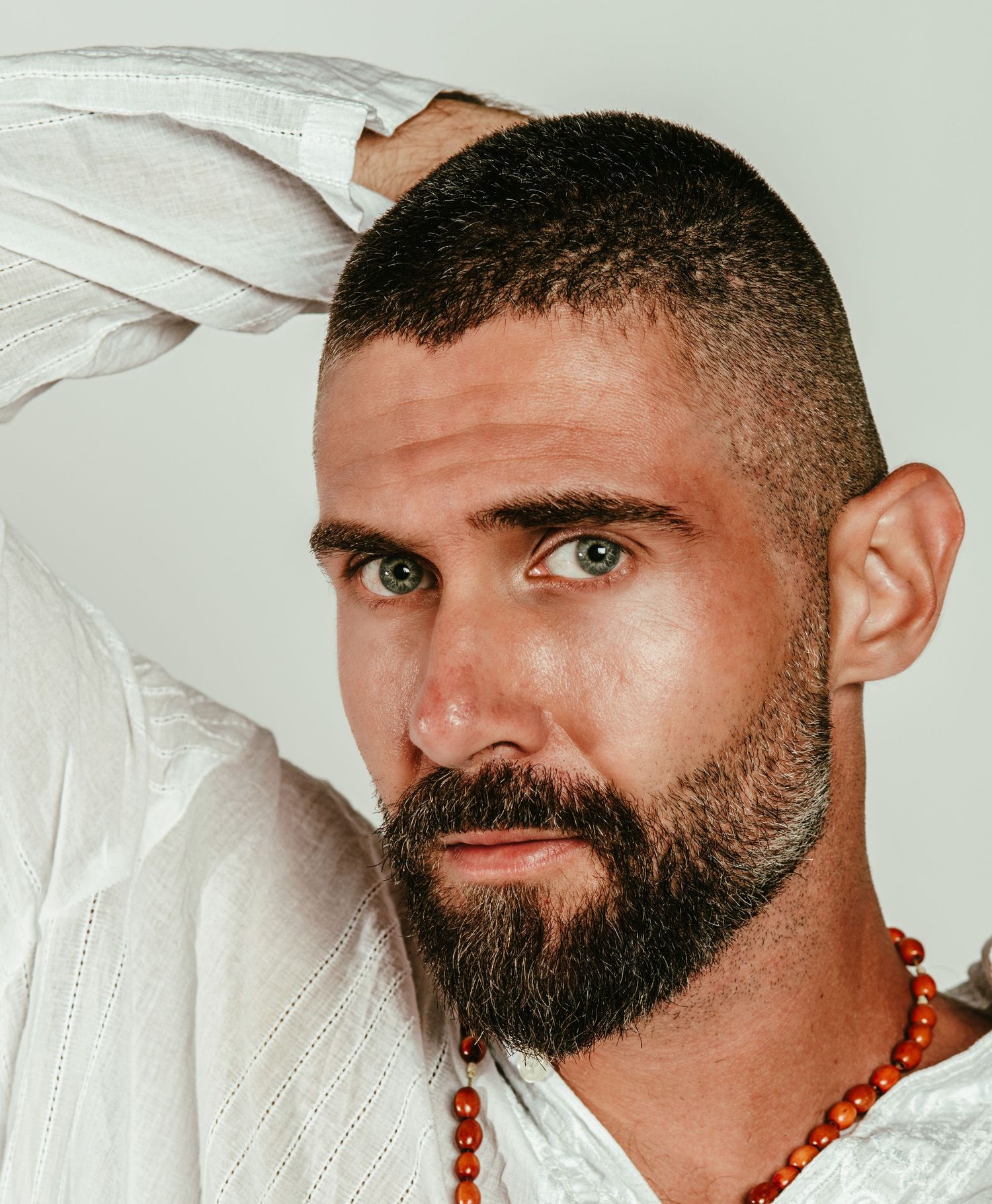 Man with short hair, beard, green eyes, white shirt, and orange beaded necklace posing against light background.