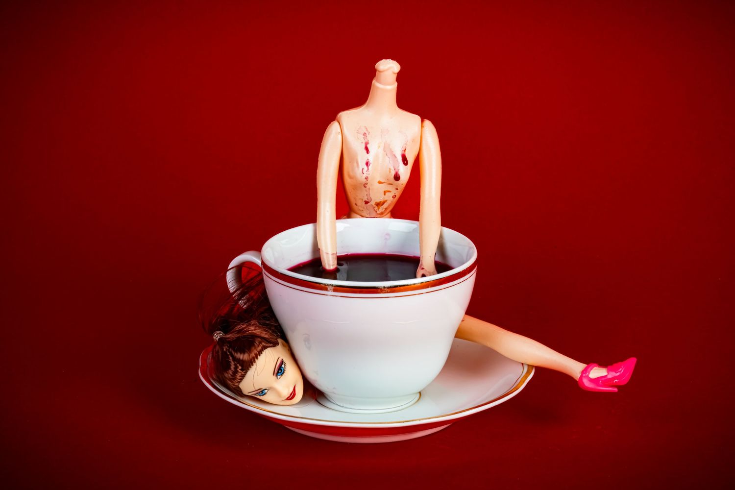 Miniature doll figure lounging in white teacup against dramatic red background.