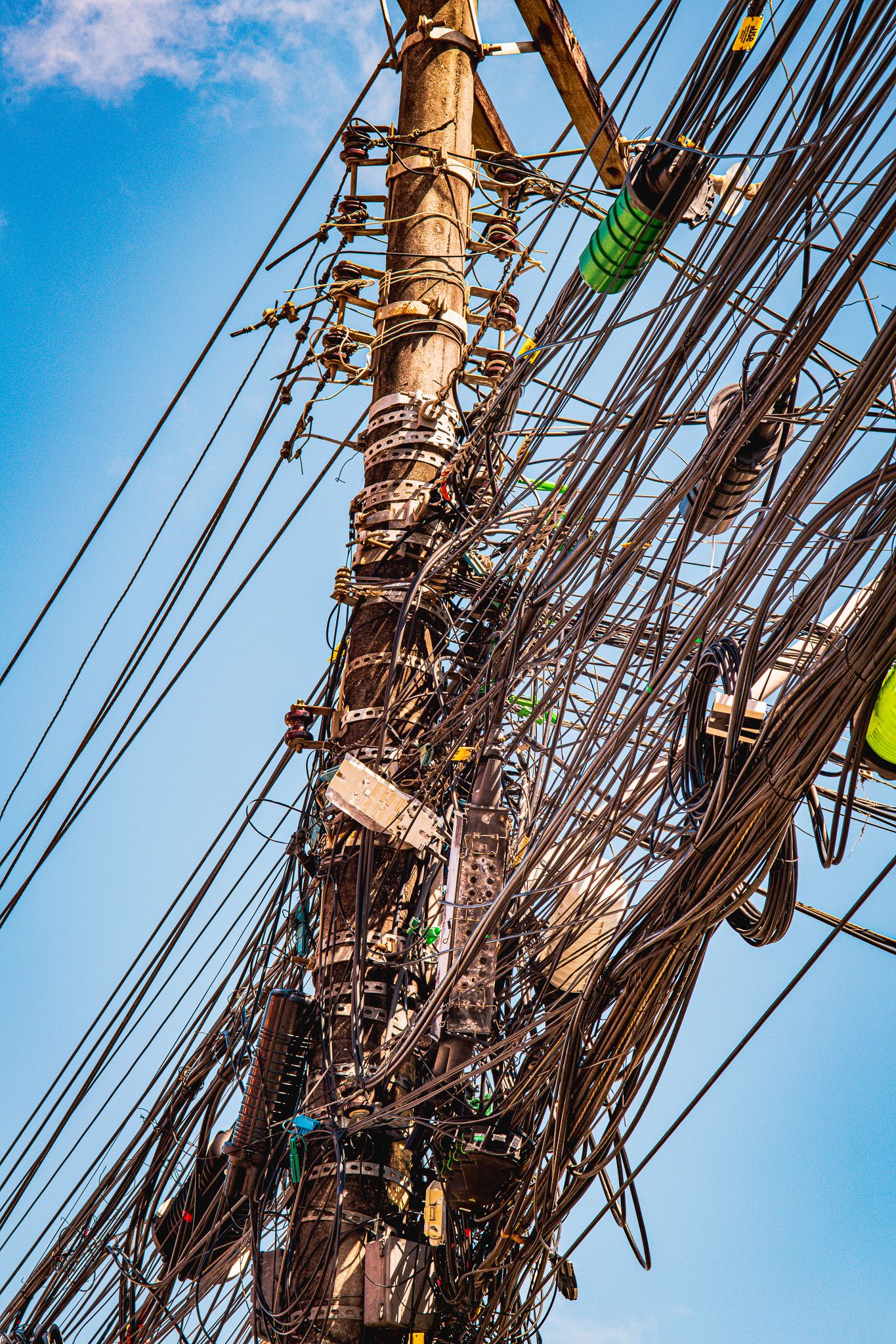 Overcrowded utility pole with dense maze of power lines and transformers against blue sky.