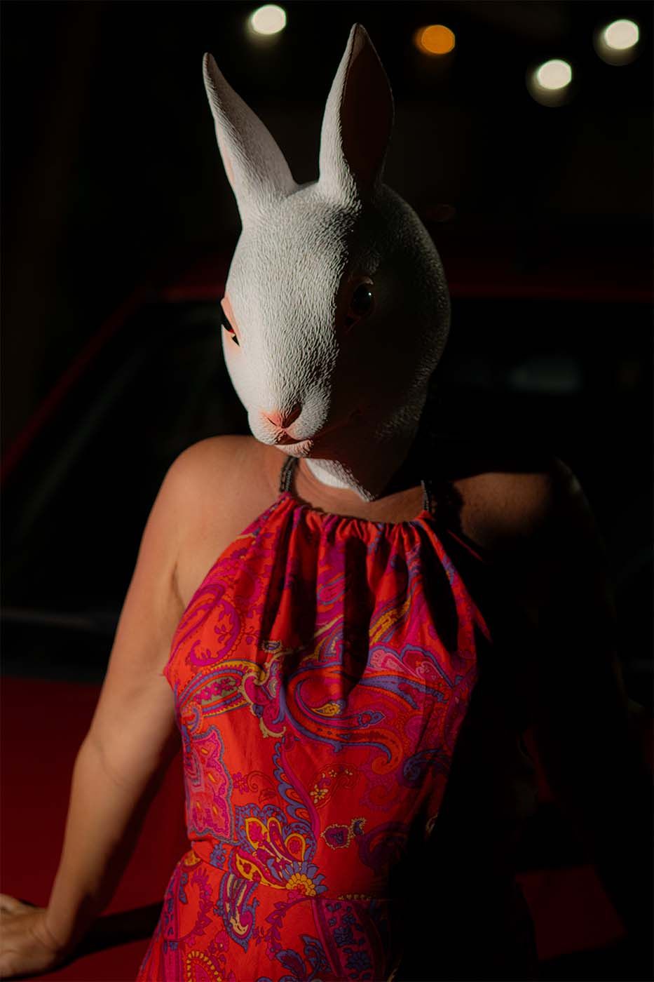 Person wearing a white rabbit mask and vibrant red paisley halter dress standing near a red car at night.
