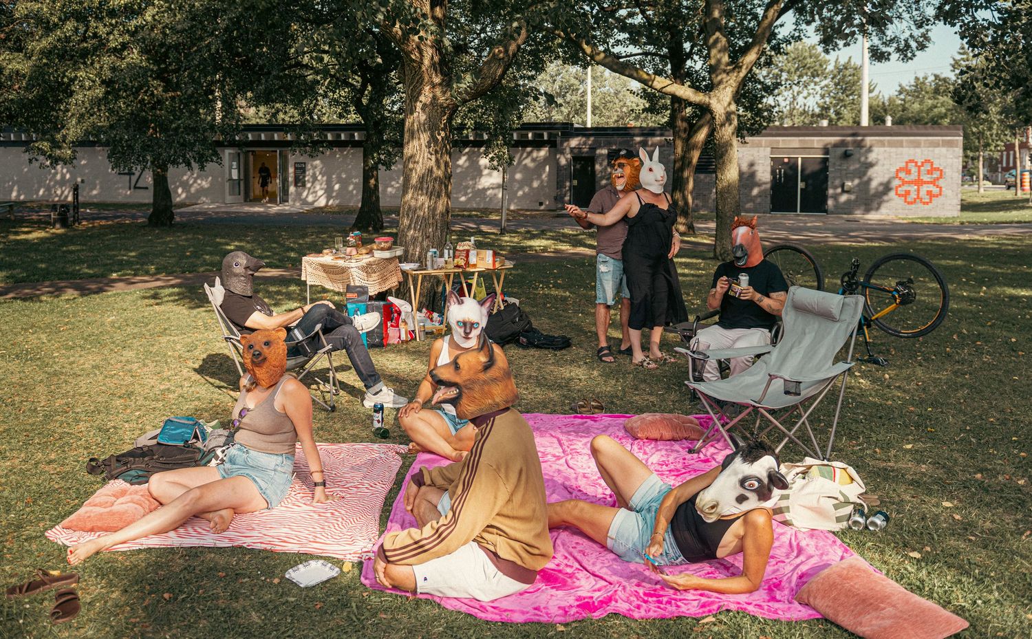 Group of people wearing animal masks enjoying a sunny outdoor picnic on colorful blankets in a park.