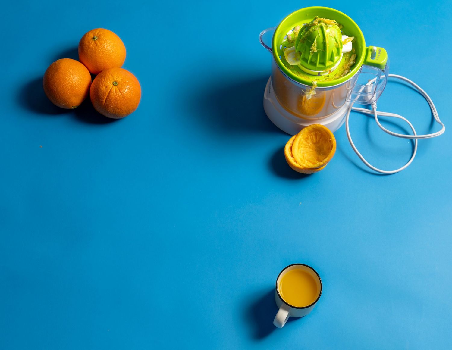 Fresh oranges beside a green electric citrus juicer and a cup of orange juice on a blue background.