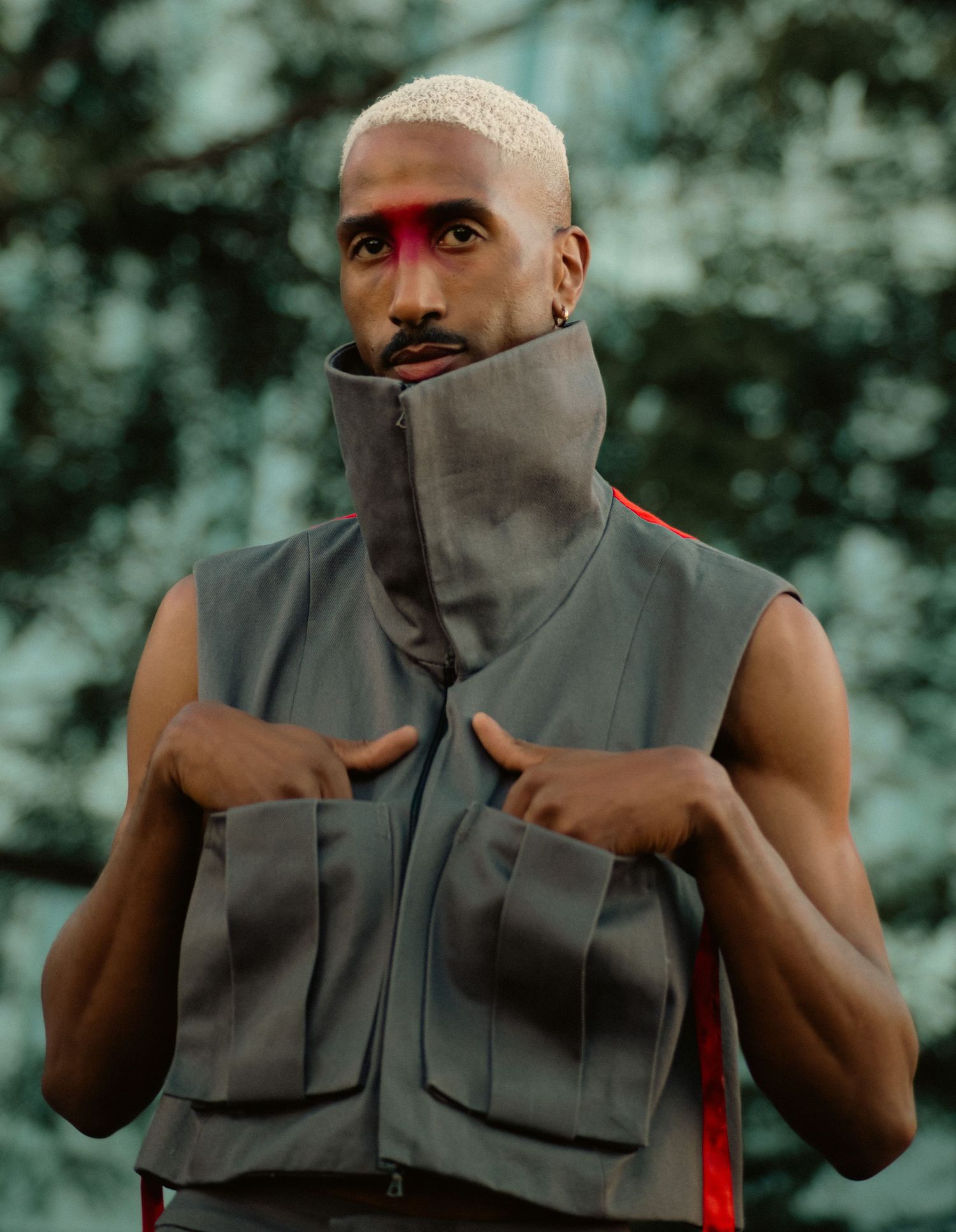 A muscular man with bleached hair and red face paint wears a gray tactical vest outdoors.