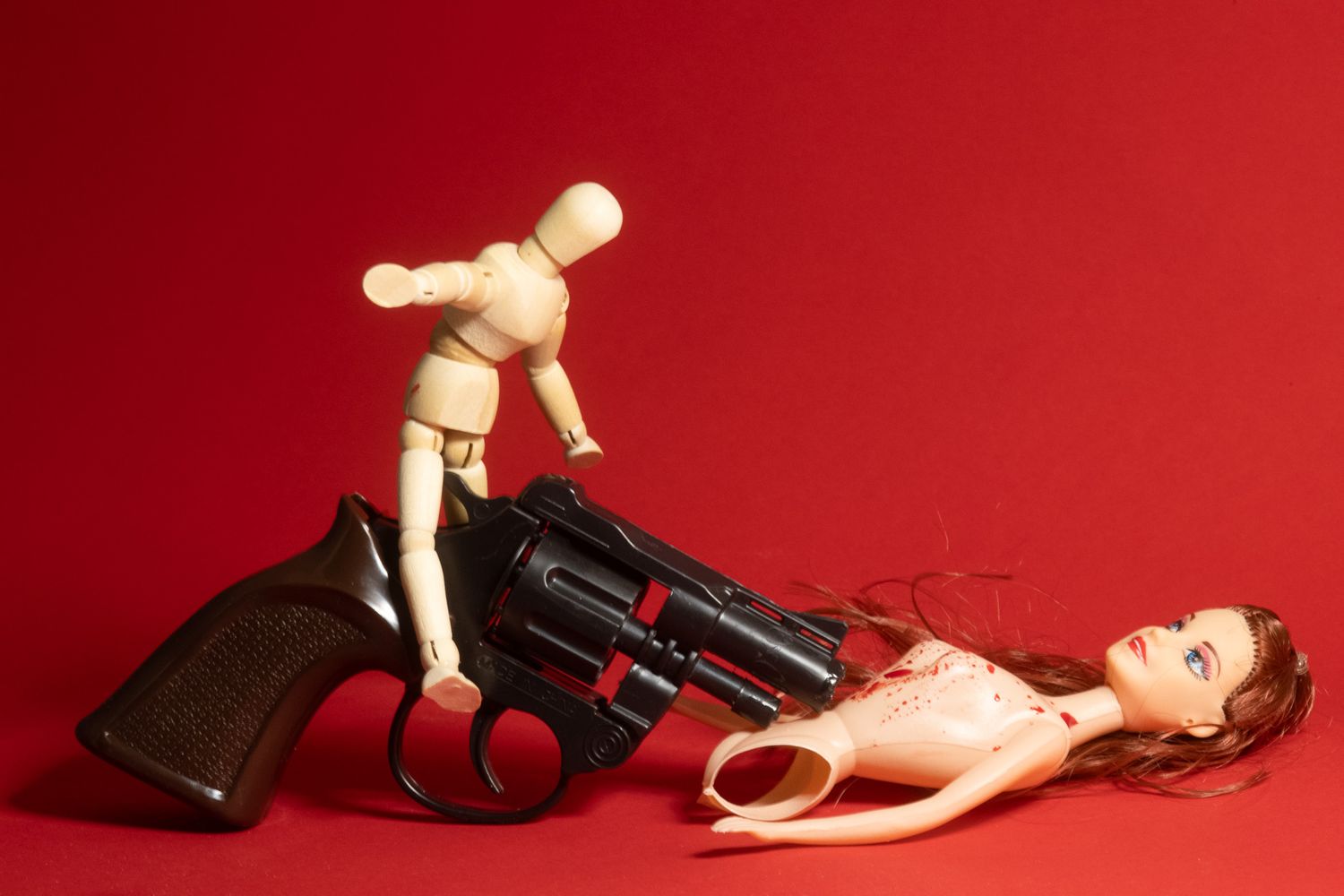 Wooden artist mannequin holding toy gun against red background in dramatic lighting.