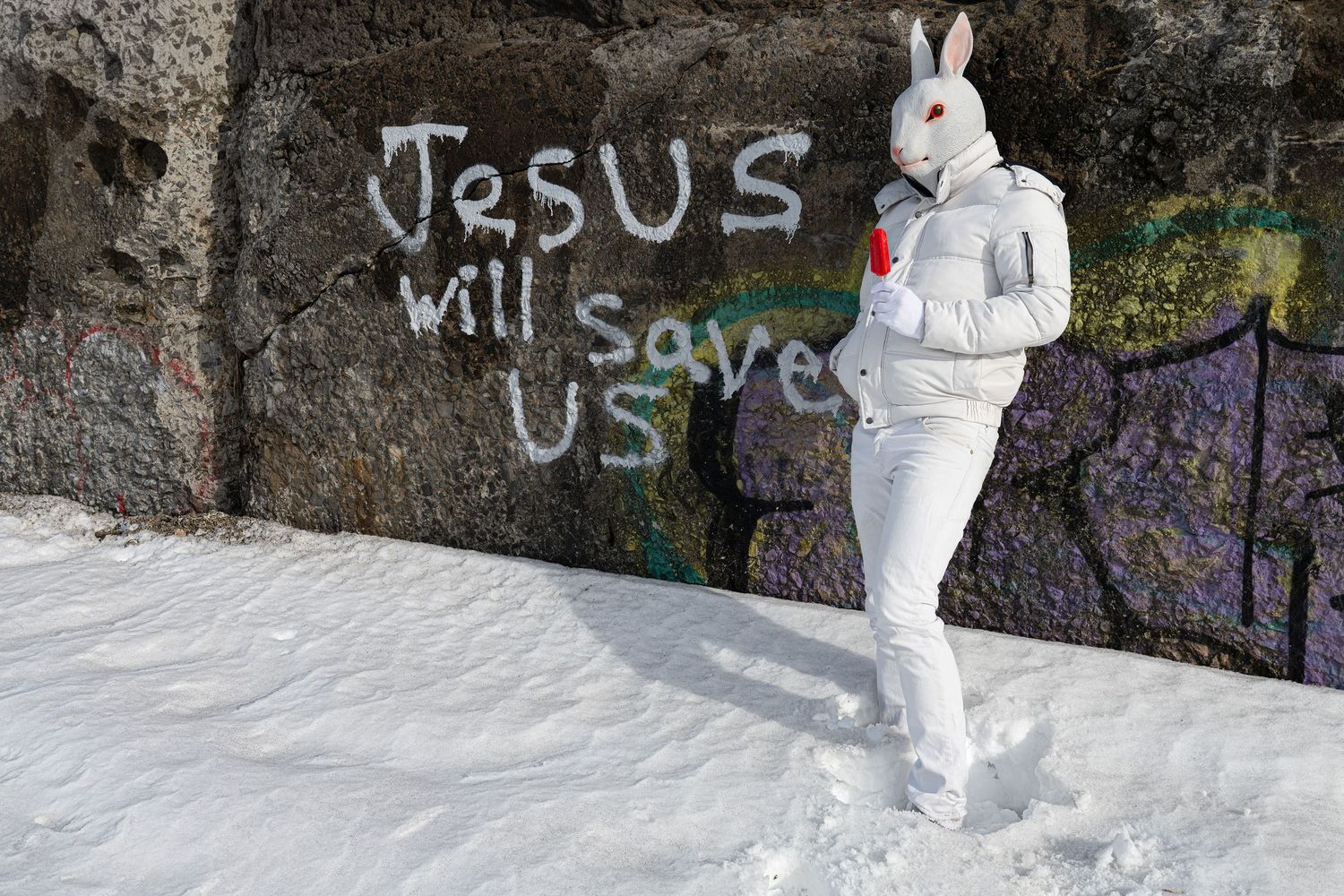 Person in white bunny costume standing in snow near graffiti reading Jesus Will Save Us on a stone wall.