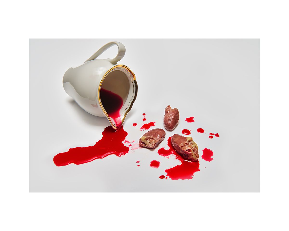 Overturned white and gold cup spilling bright red liquid with scattered chocolate candies on white surface.