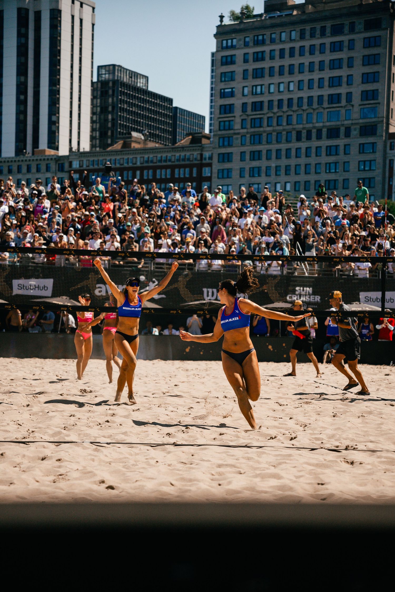 Beach volleyball players competing on sandy court with city skyline backdrop.