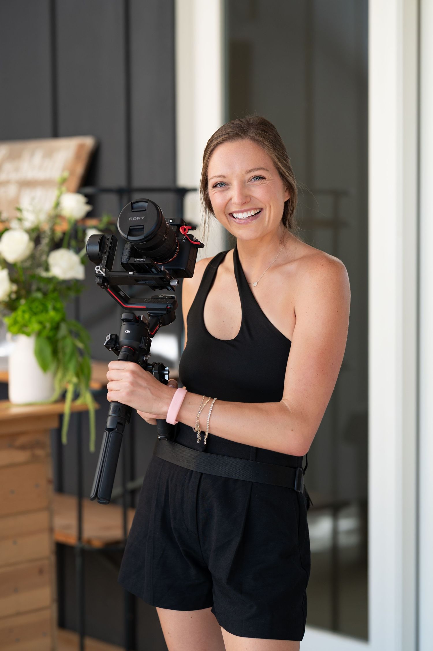 Professional photographer wearing black dress holding camera and smiling in studio setting.