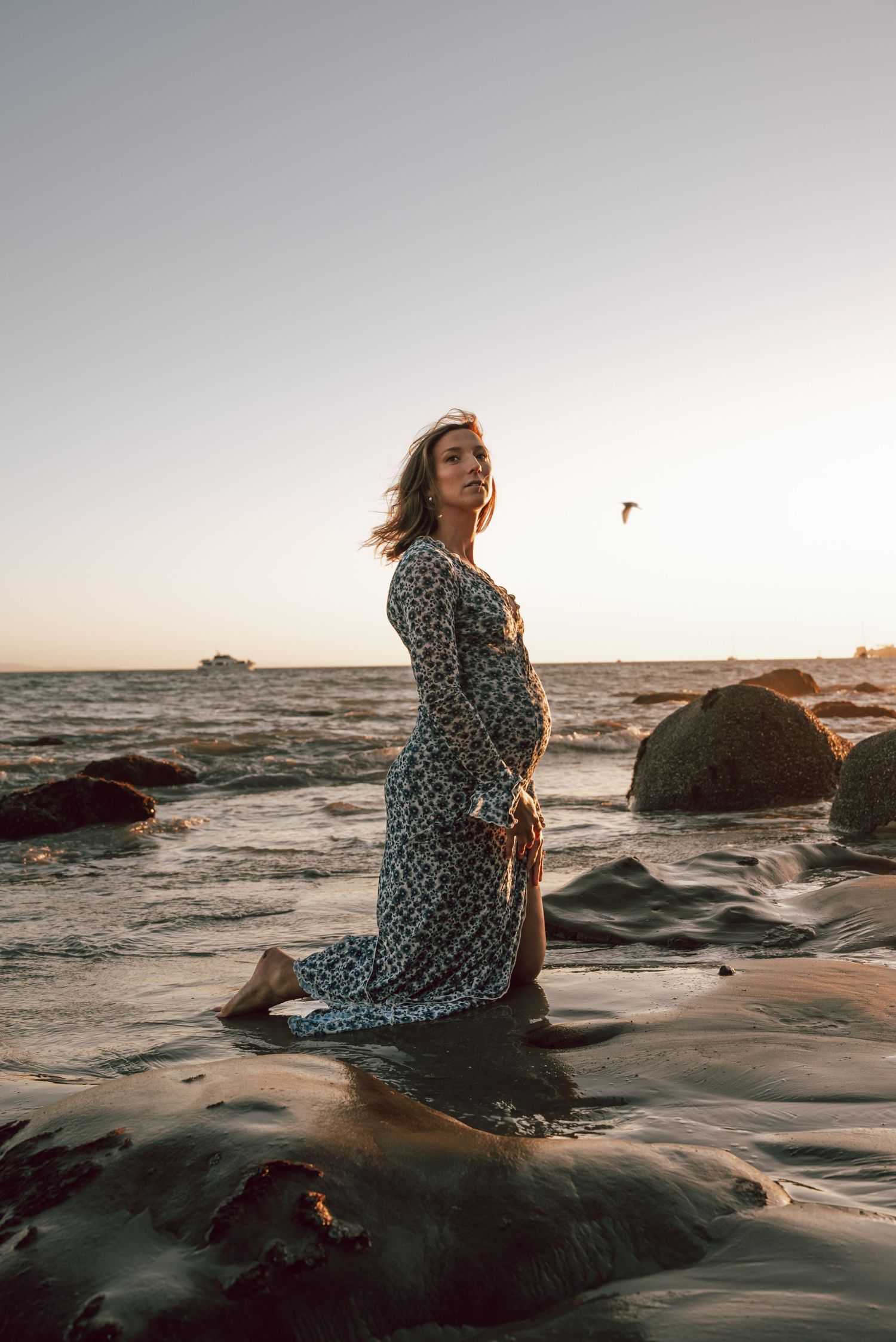 Woman in floral dress poses on rocky beach during sunset.