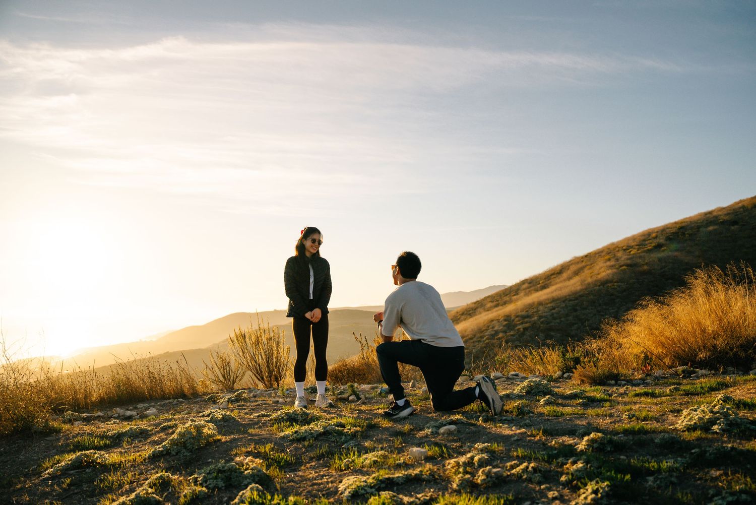 Two people sit together on a grassy hillside at sunset enjoying scenic mountain views.