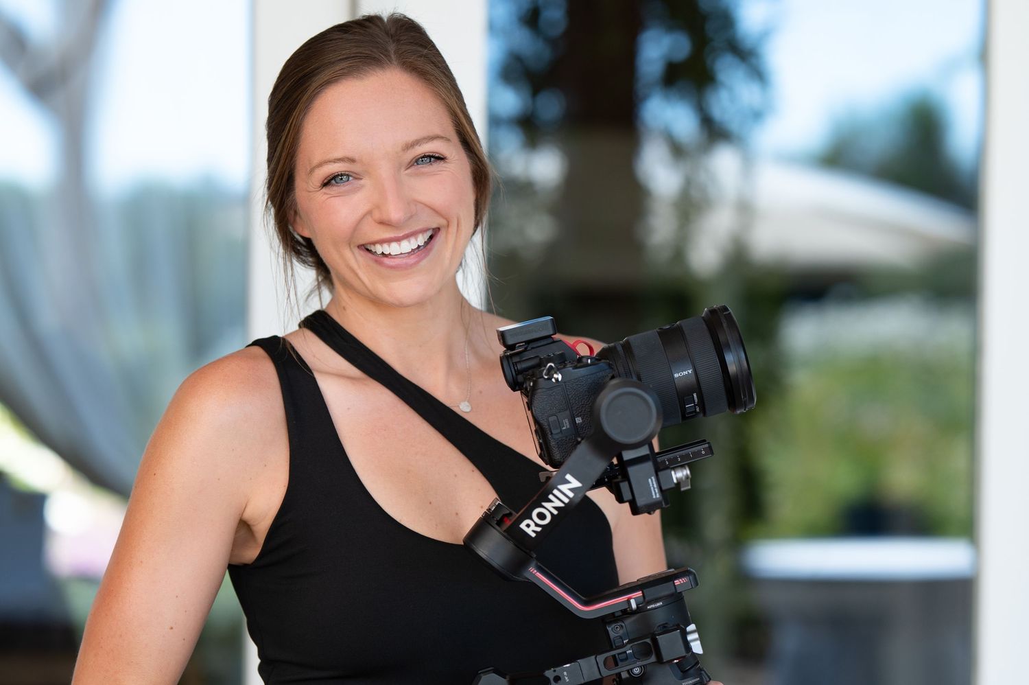 Woman in black outfit poses with professional camera equipment against bright background.