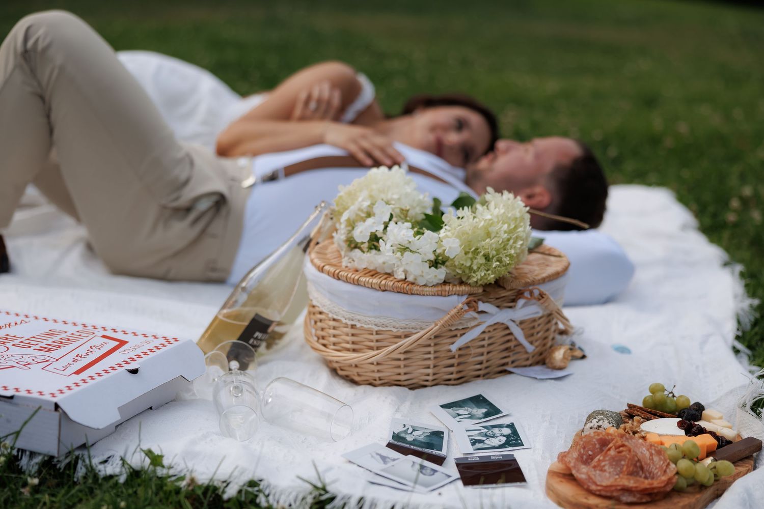 Picnic spread with basket, flowers and snacks on white blanket in garden.