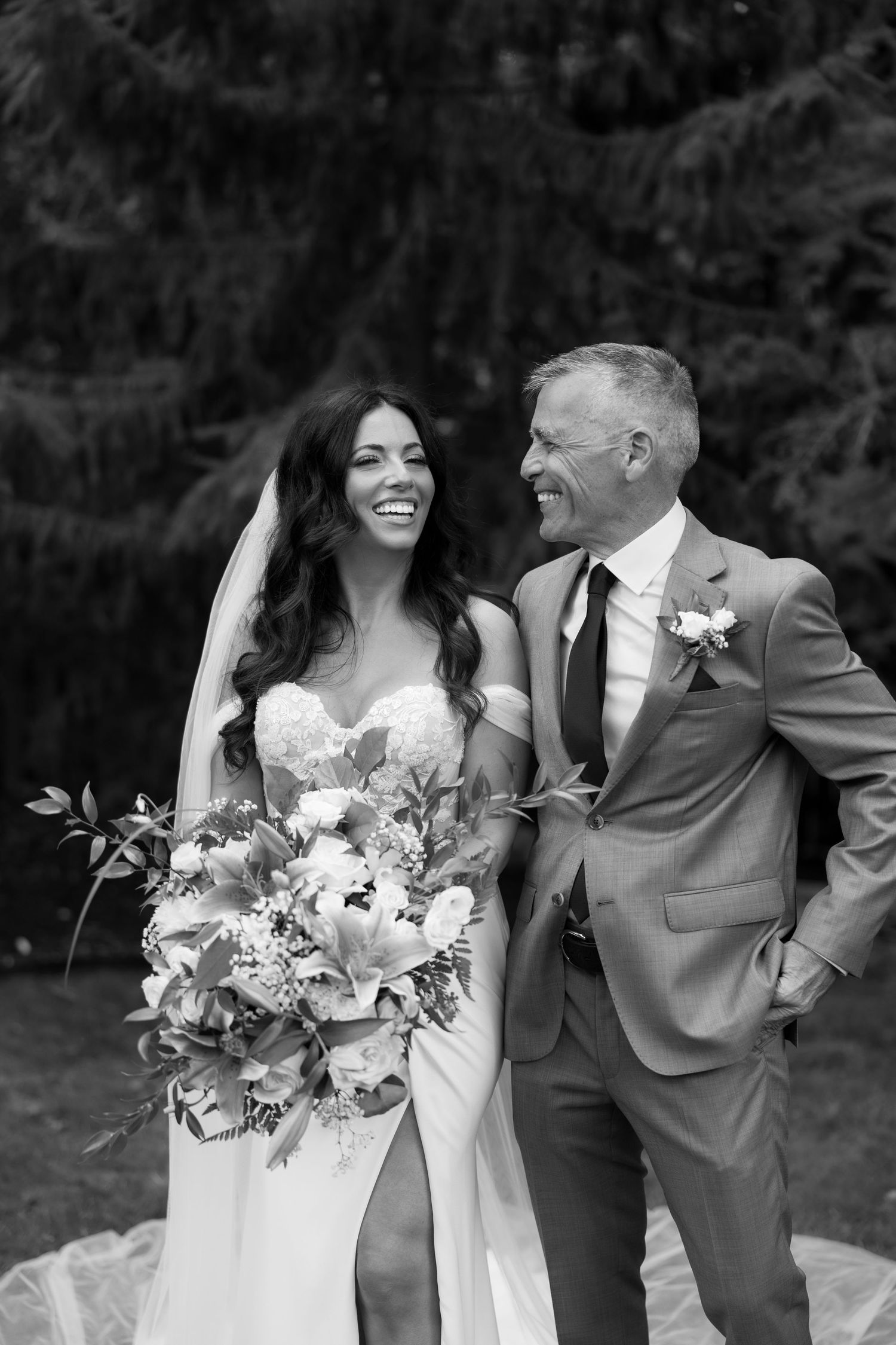 Joyful wedding couple laughing while holding floral bouquets outdoors.