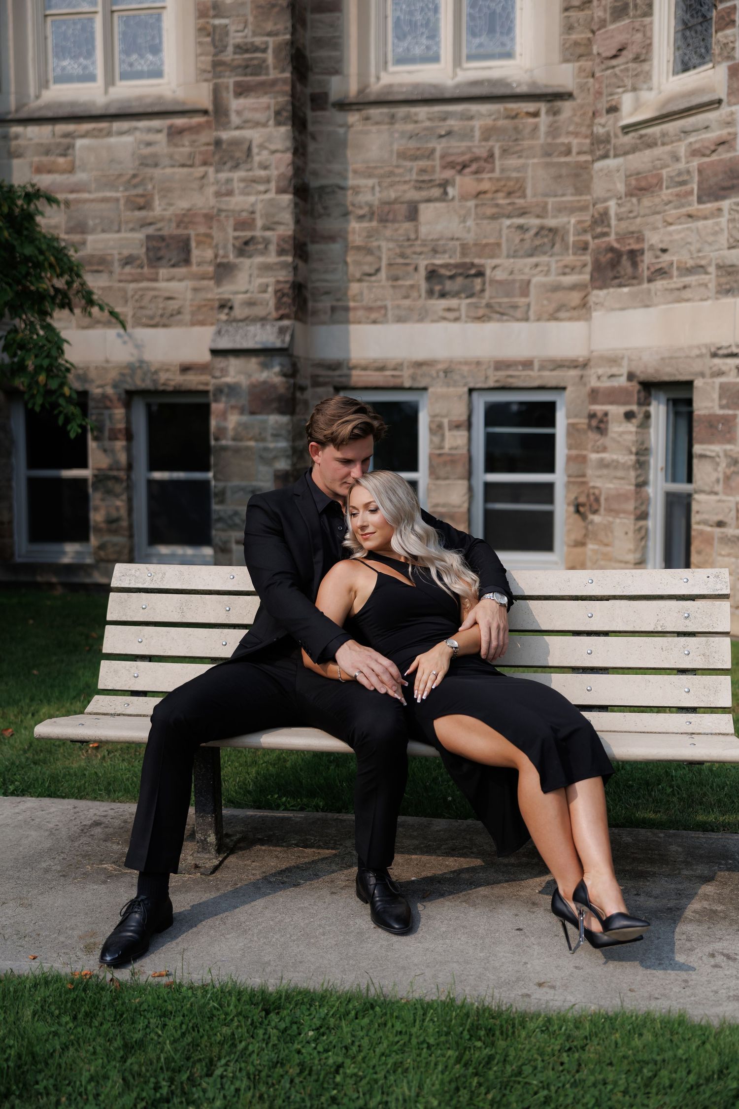 Couple in black attire posing together on outdoor bench near stone building.