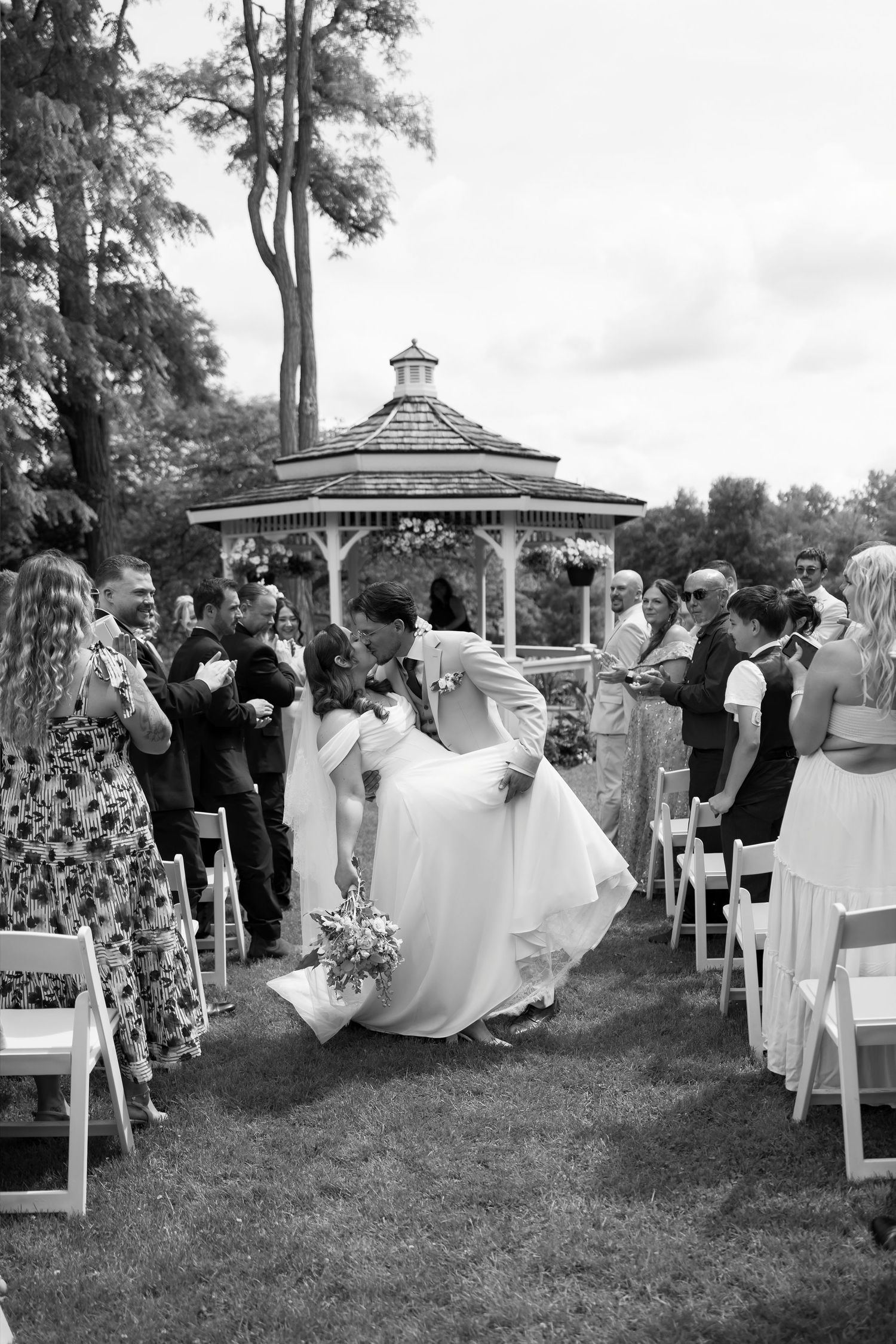 Black and white photo of bride and groom sharing a dip kiss while walking down the aisle at an outdoor gazebo wedding ceremony.