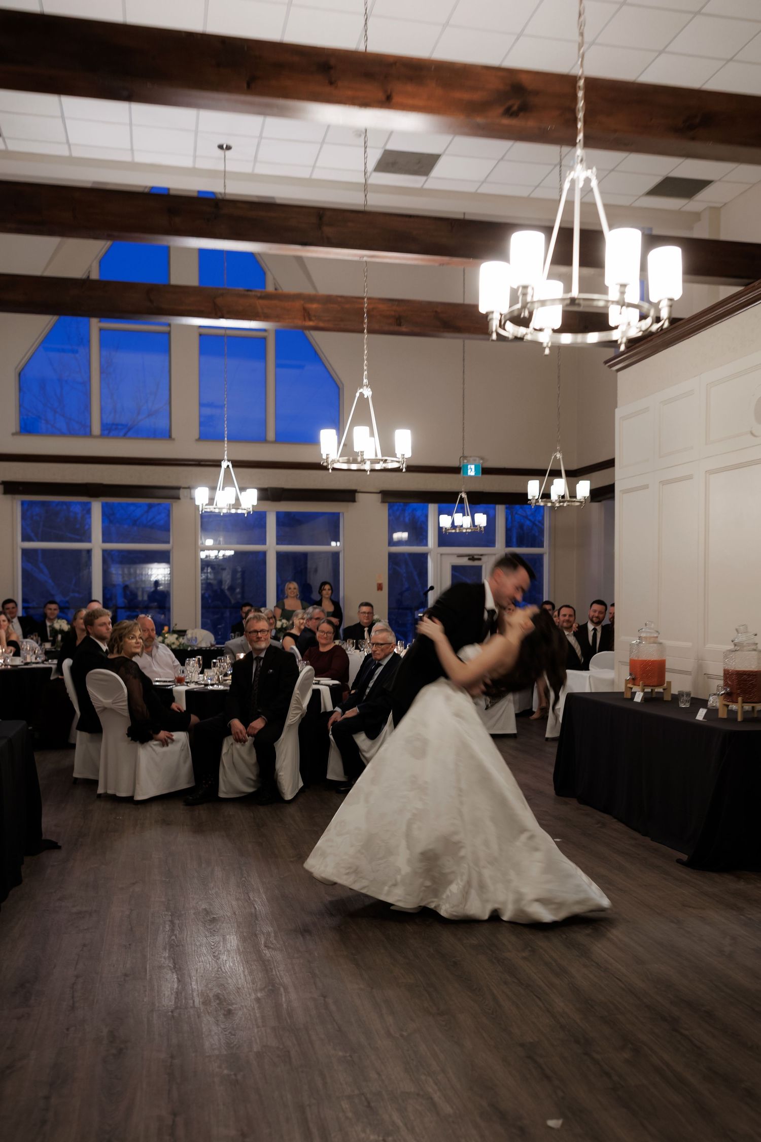Bride and groom share first dance in elegant reception hall with chandeliers and wooden beam ceiling.