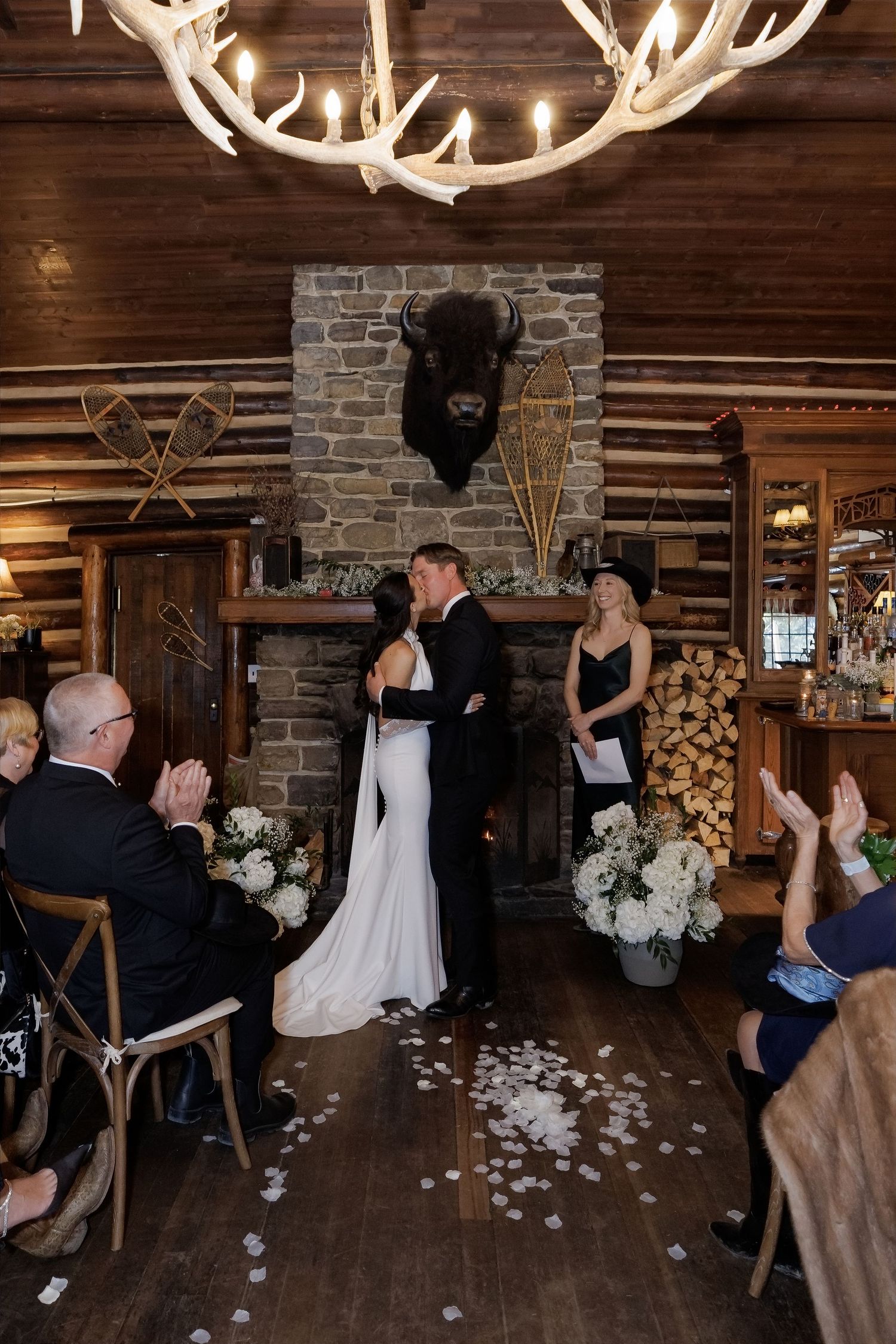 Couple shares first kiss at rustic log cabin wedding ceremony with stone fireplace and bison mount decor.