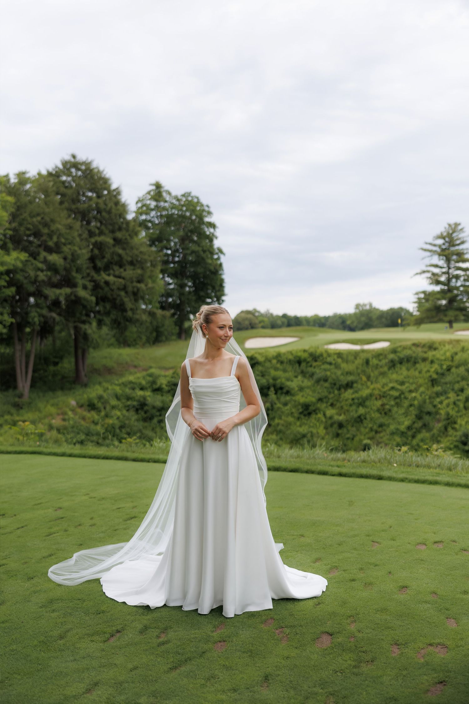 Elegant bride in a simple white A-line gown with cathedral veil posing on a lush green golf course lawn.