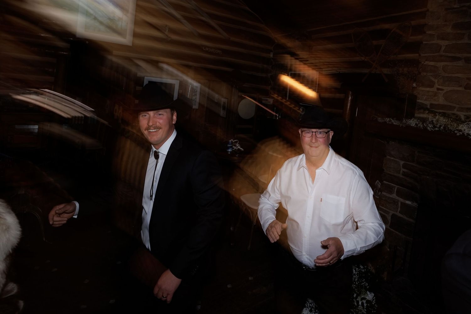 Two men wearing black cowboy hats and dress shirts stand in a dimly lit rustic interior.